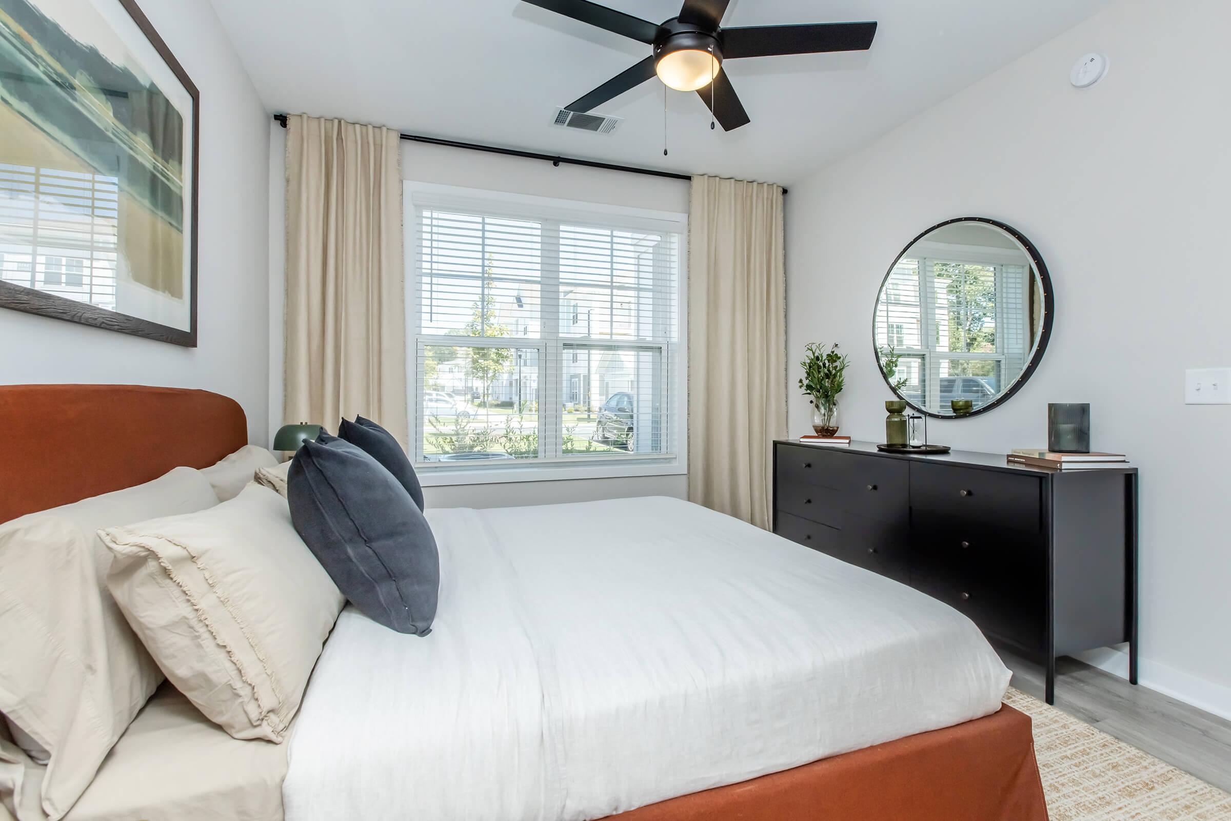 A cozy bedroom featuring a bed with neutral bedding and decorative pillows, a stylish black dresser, a round mirror, and large windows with white curtains letting in natural light. A small plant adds a touch of greenery to the space, enhancing the serene atmosphere.
