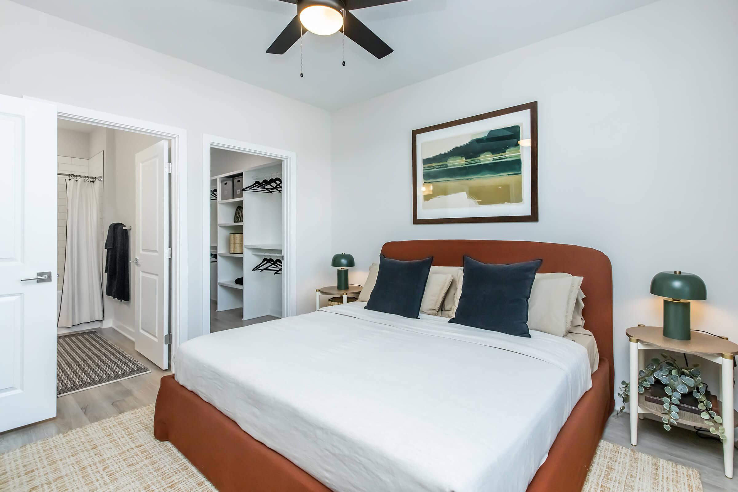 A cozy bedroom featuring a brown upholstered bed with neutral bedding and dark blue pillows. There's a painting on the wall, and a bedside table with a green lamp. A closet is visible on the left with sliding doors, and a light rug covers the floor.