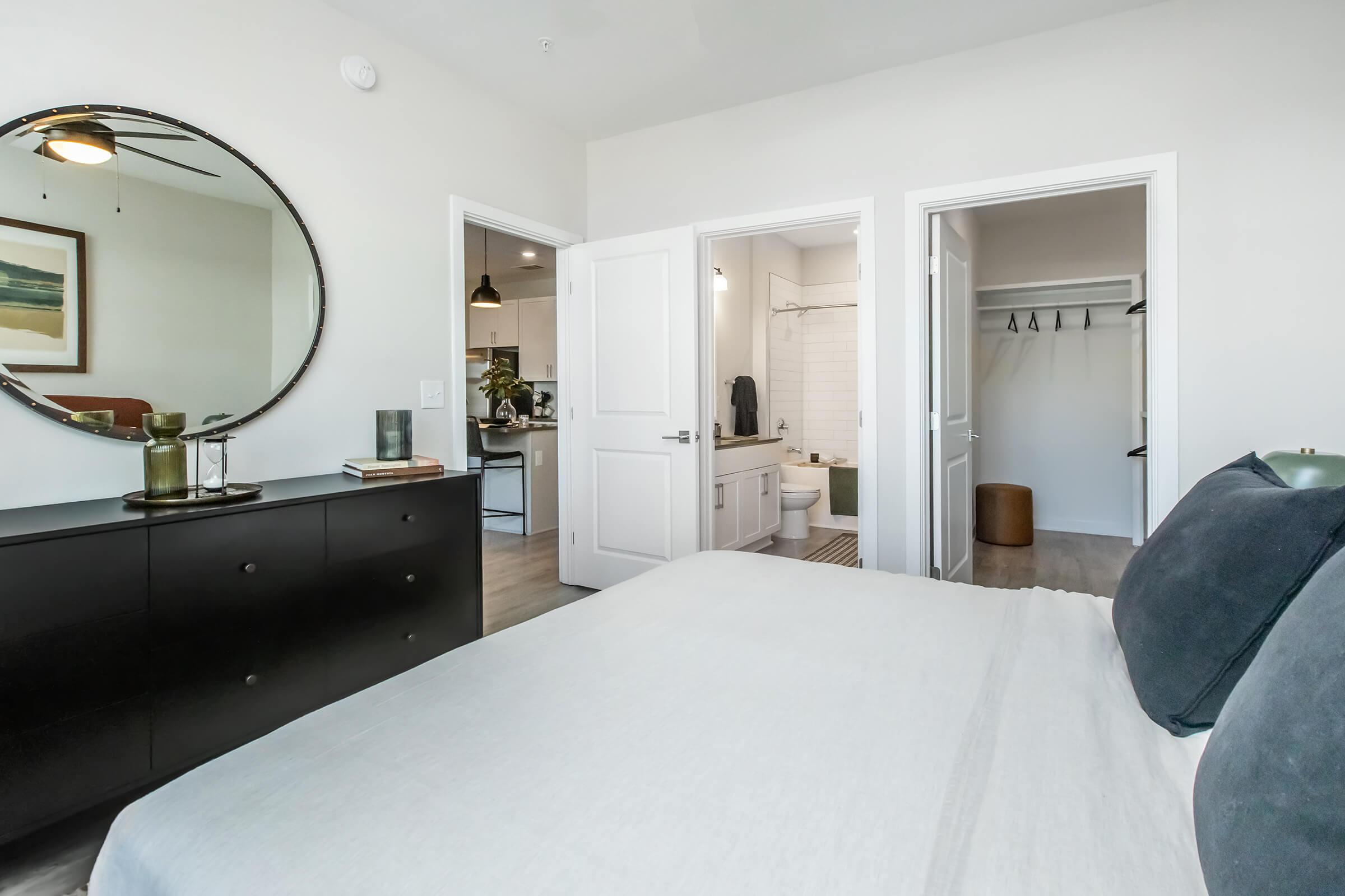 A modern bedroom featuring a large round mirror above a dark dresser, with a neatly made bed in the foreground. In the background, there are two doors leading to a bathroom and a closet, with soft neutral colors and stylish decor enhancing the tranquil atmosphere.