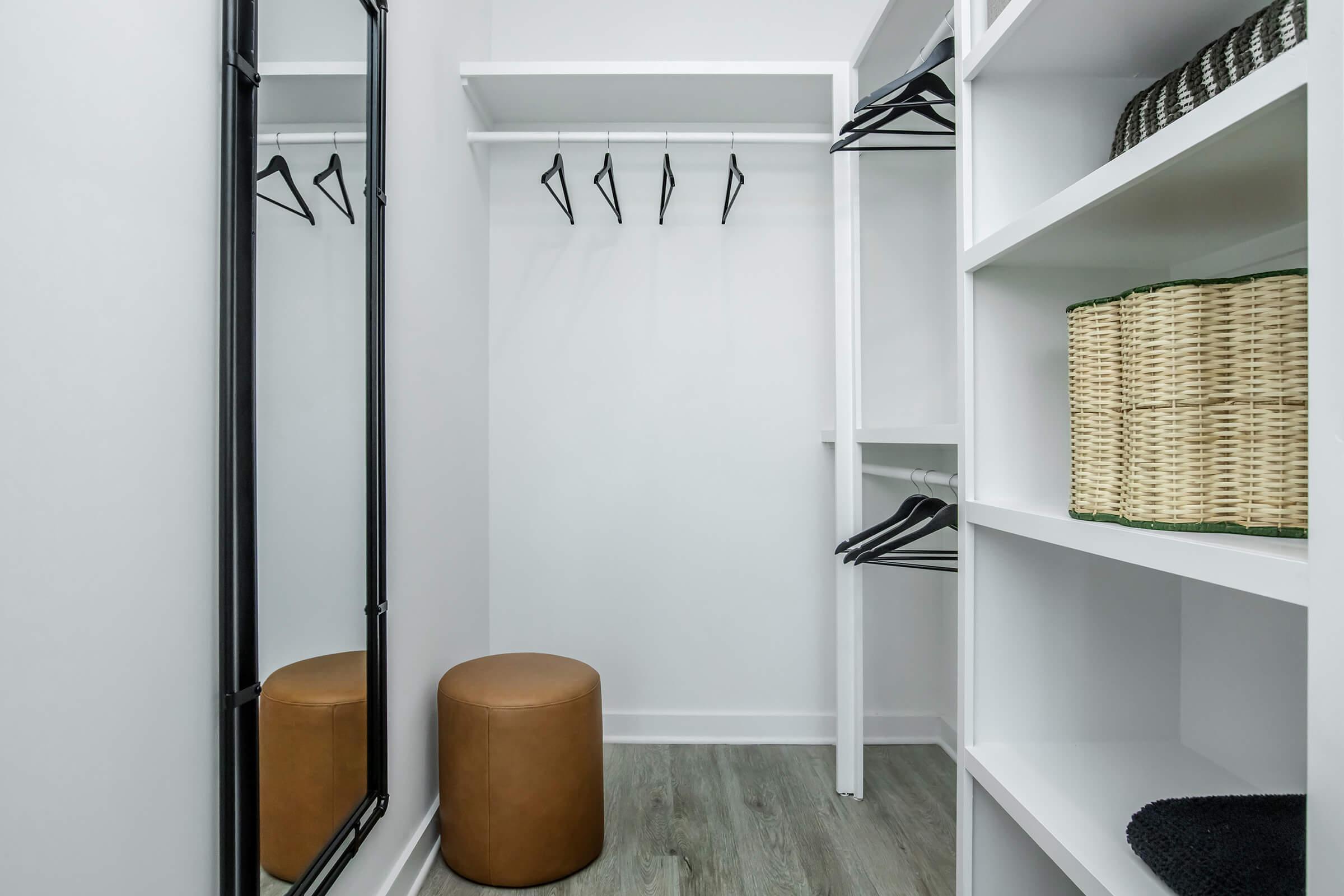 A modern, minimalist closet with a full-length mirror, black hangers on a shelf, and a tan ottoman. The shelves are white, containing woven storage baskets and a neatly arranged space, providing an organized look. The floor features light-colored wood, enhancing the clean aesthetic.