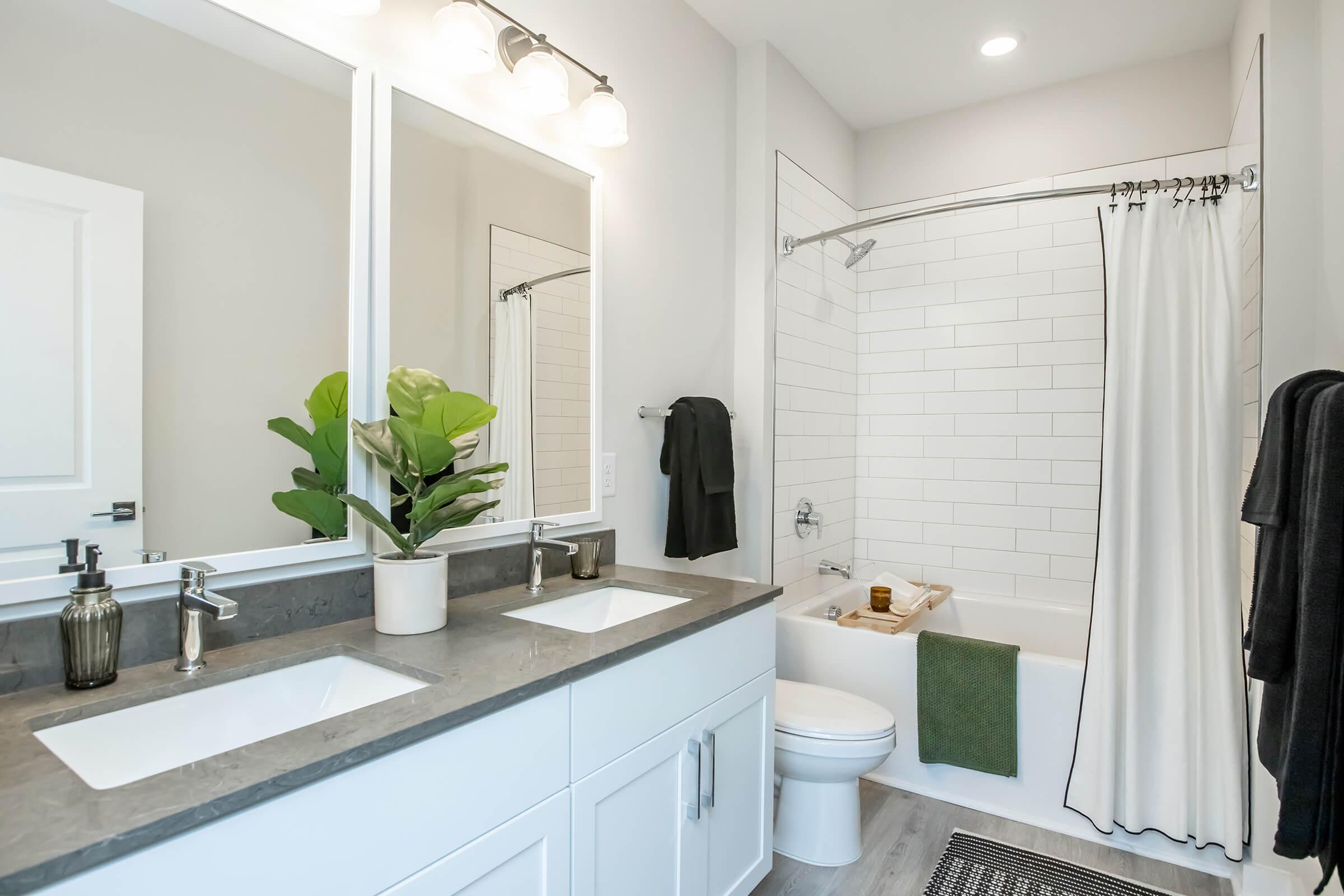 A modern bathroom featuring a double vanity with white sinks, a large mirror, a tub with a shower curtain, and light-colored walls. A small potted plant adds a touch of greenery, while bath towels hang nearby. The flooring is light wood, enhancing the fresh and clean aesthetic.
