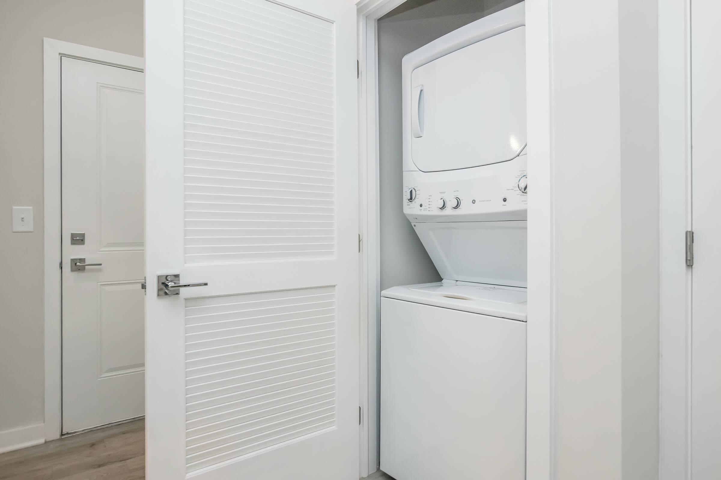 A laundry room featuring a stacked washer and dryer unit, partially visible behind a white door. The room has light gray walls and a modern, clean aesthetic, with a second door and wooden flooring in the background.