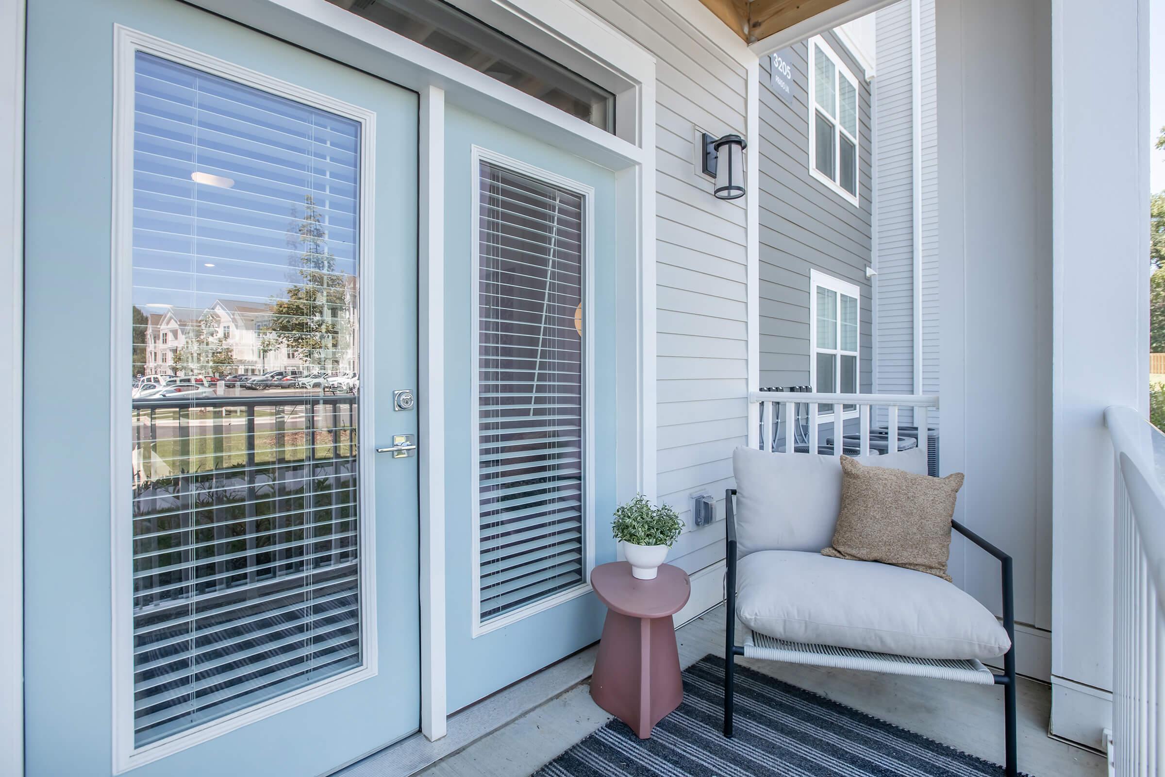 A cozy outdoor space featuring a light blue entrance door with glass panels, a small round table, a comfortable chair with a cushion, and a potted plant. The area is adorned with a striped rug and has a view of a neighboring building and greenery. Natural light streams in, creating a welcoming atmosphere.