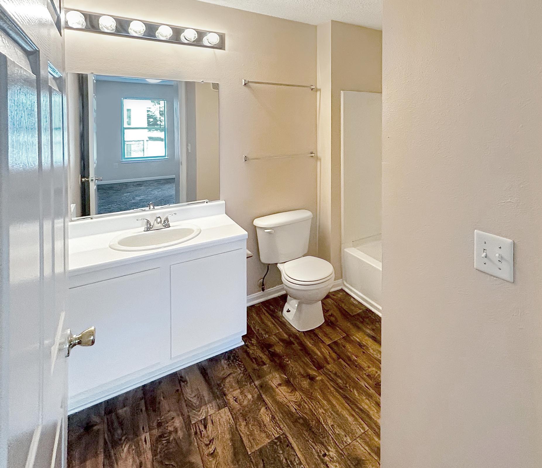 A bright bathroom featuring a white vanity with a sink, a mirror, a toilet, and a bathtub. The floor is finished with wood-like tiles, and the walls are painted a light beige. Natural light enters from a nearby window in the background, creating an inviting atmosphere.