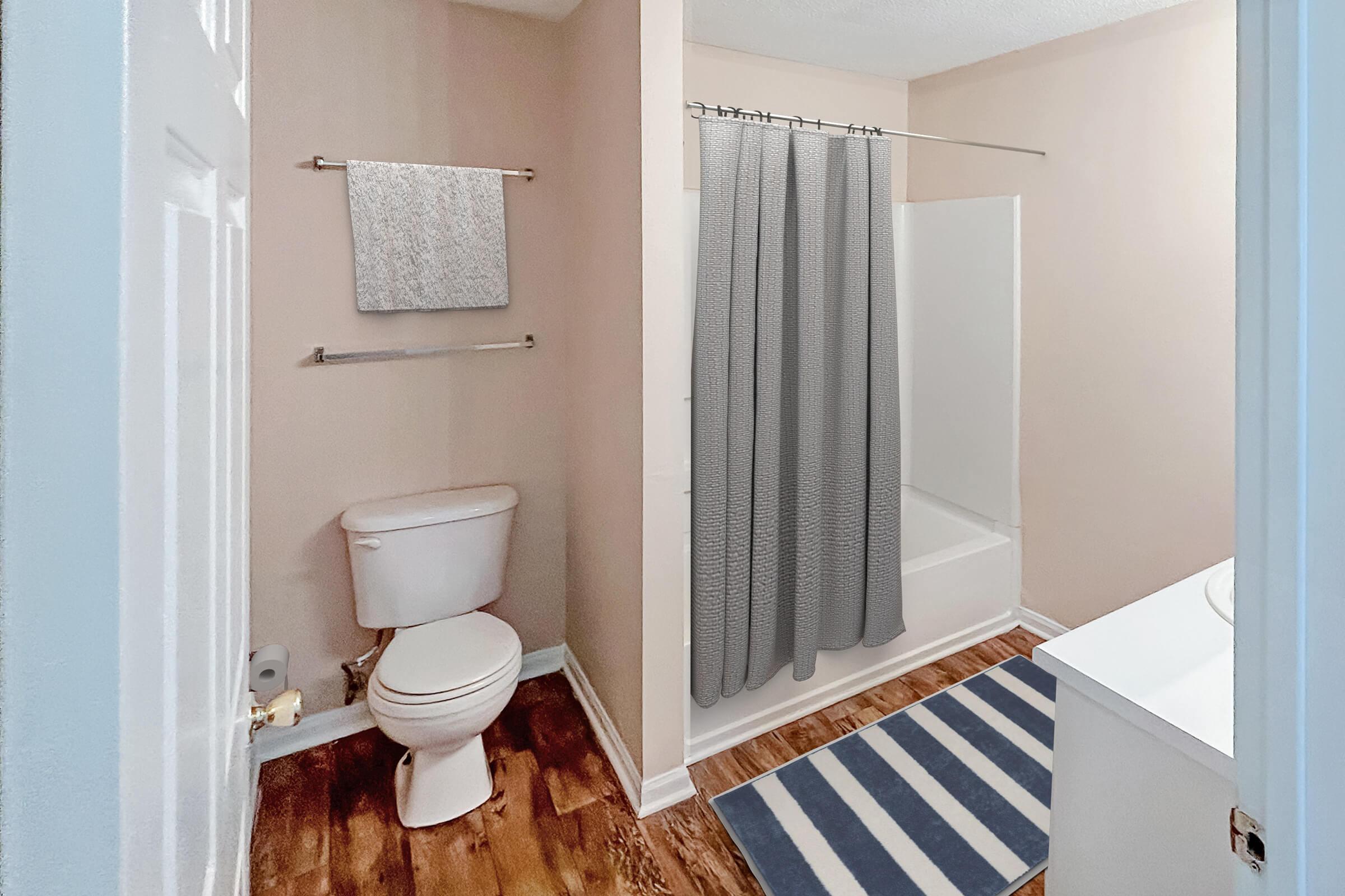 A clean and modern bathroom featuring a white toilet, a bathtub with a gray shower curtain, and a wall-mounted towel rack. The floor has a wood-like finish, and a blue and white striped bath mat is positioned in front of the bathtub. Light-colored walls enhance the bright and spacious feel of the room.