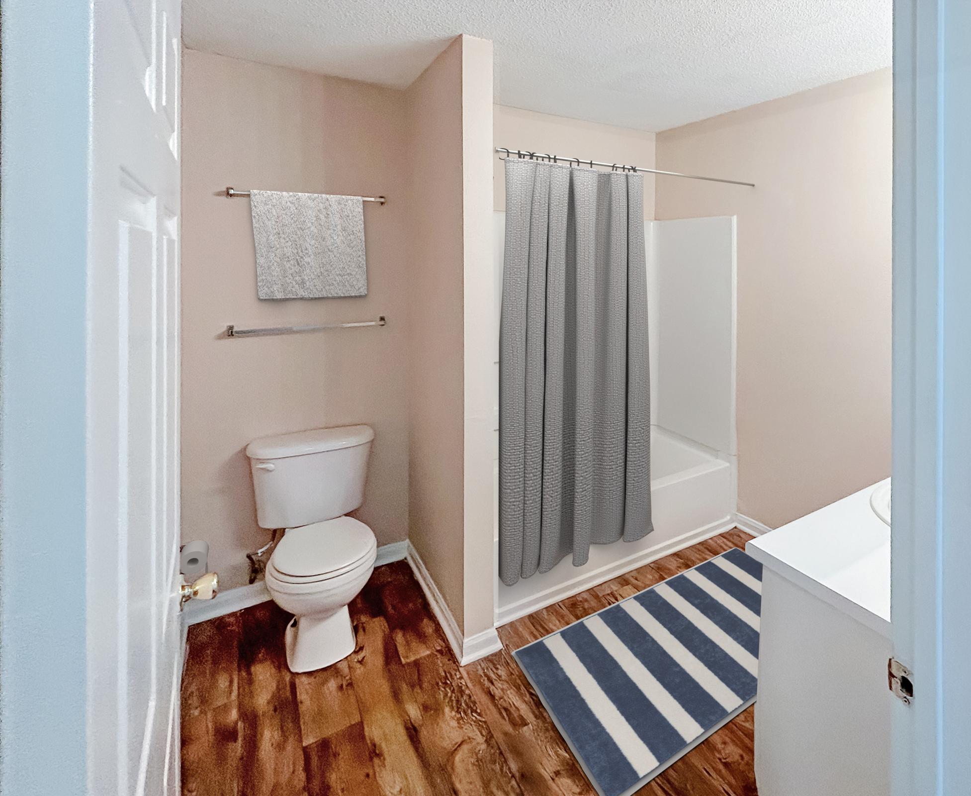 A clean and minimalistic bathroom featuring a toilet, a shower with a gray curtain, a white bathtub, and a light-colored wall. The floor has a hardwood appearance, complemented by a blue and white striped rug in front of the sink area. A gray towel hangs on the wall.