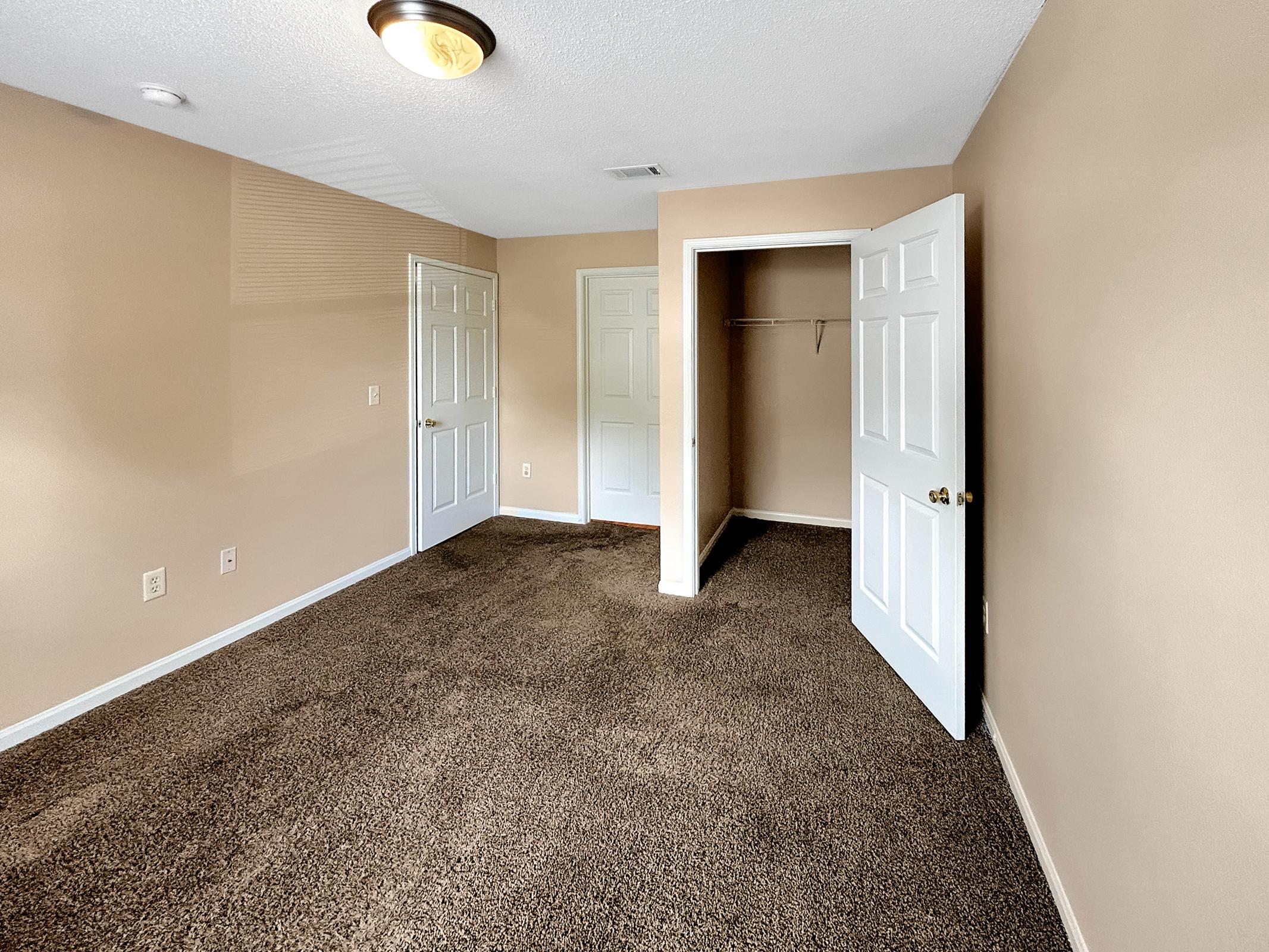 A spacious, empty room with beige walls and brown carpeting. Two closed white doors are visible, one leading to a closet with a single hanging rod. Natural light fills the space, highlighting the minimalist design and clean, open layout.