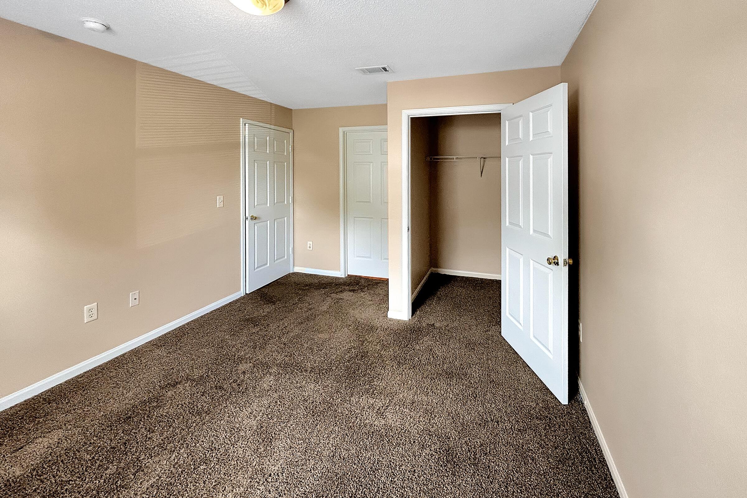 Empty room with beige walls and carpeted floor. There are two white doors visible, one leading to a closet. The space is well-lit and appears to be freshly painted, creating a simple and inviting atmosphere.
