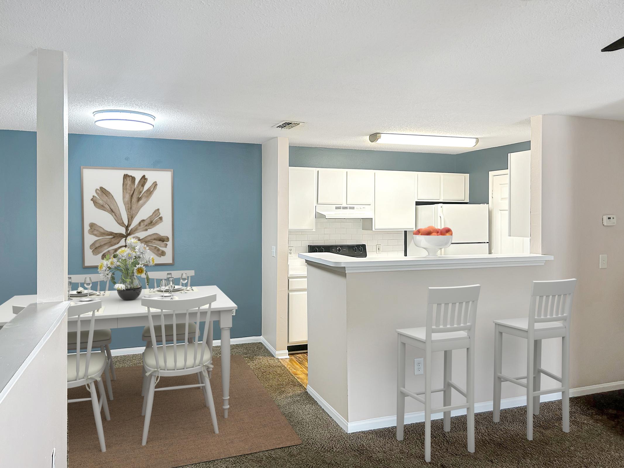 A modern kitchen and dining area featuring a white table set for four, beige chairs, and a bowl of fruit. The walls are painted in light blue and beige, with a piece of art on the wall depicting a leaf design. The kitchen includes white cabinets and appliances. Soft lighting enhances the inviting atmosphere.