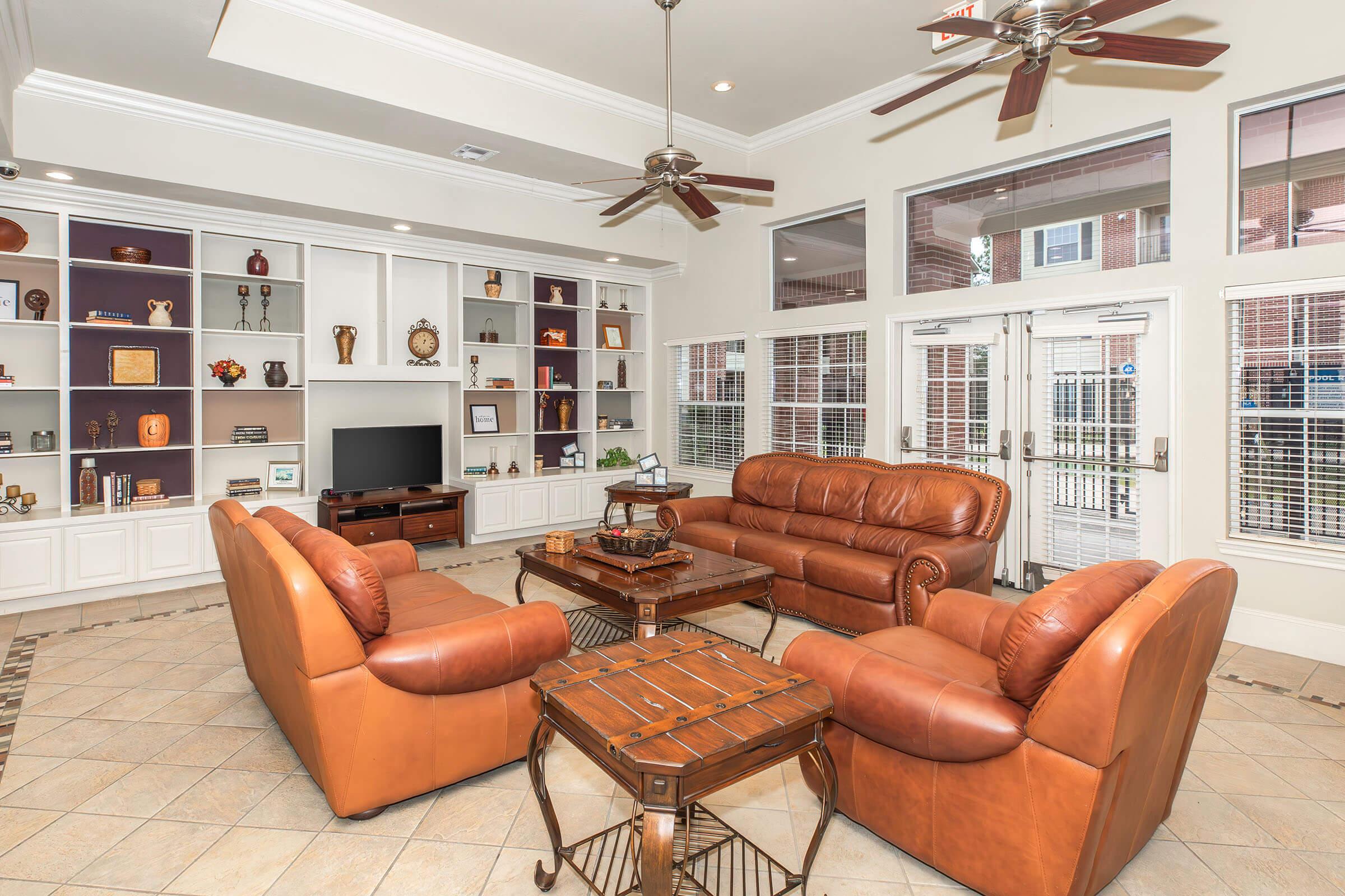 A spacious and inviting living room with a high ceiling, featuring a cozy arrangement of two leather sofas and a wooden coffee table. Built-in shelves display decorative items. Large windows allow natural light, and double doors lead to an outside area. Fans are mounted on the ceiling for comfort.