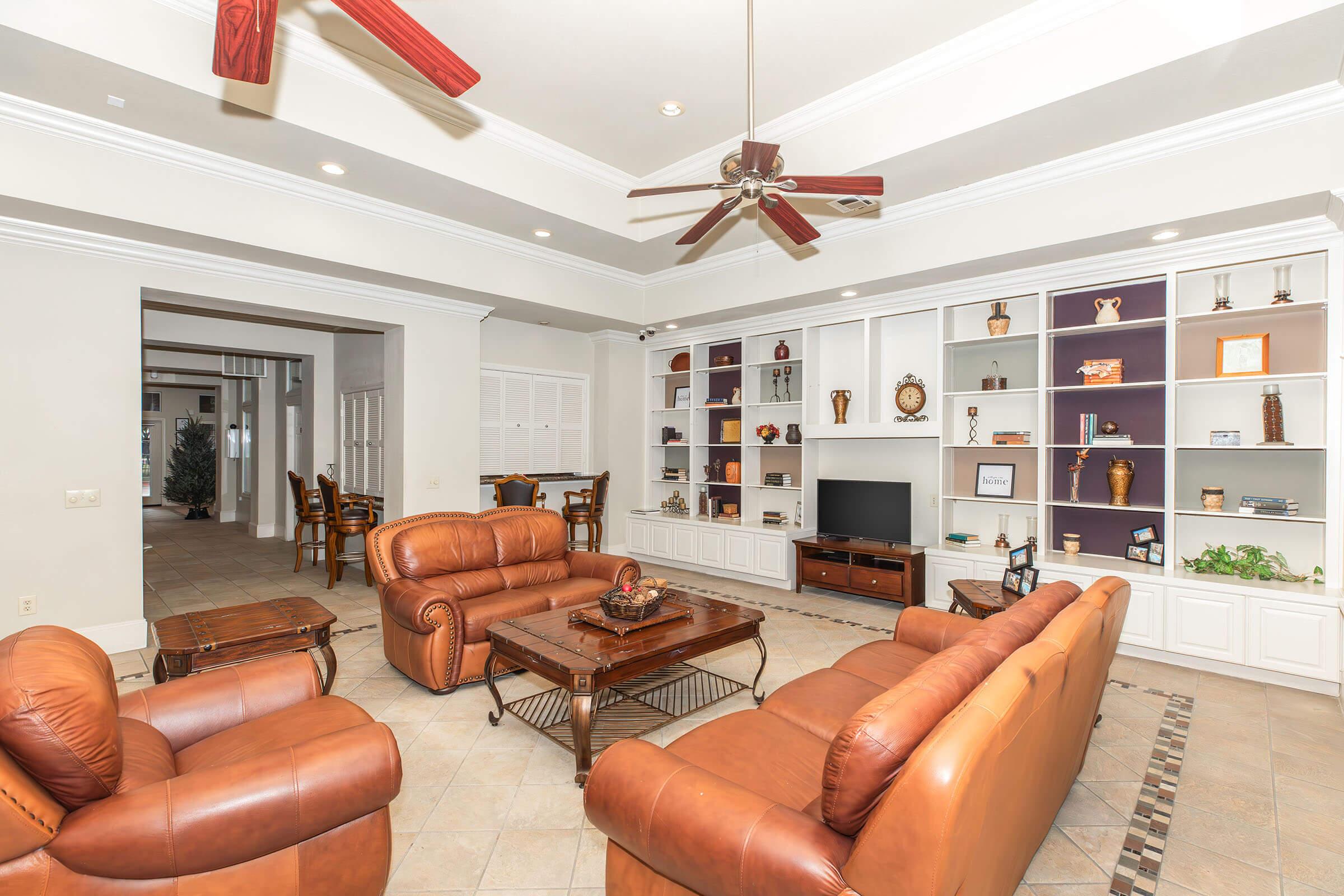 A spacious living room featuring light-colored walls, a ceiling fan, two brown leather sofas, a wooden coffee table, and built-in shelves with decorative items. A television is placed on a cabinet, and a dining area is visible in the background through an open archway. Natural light fills the space.