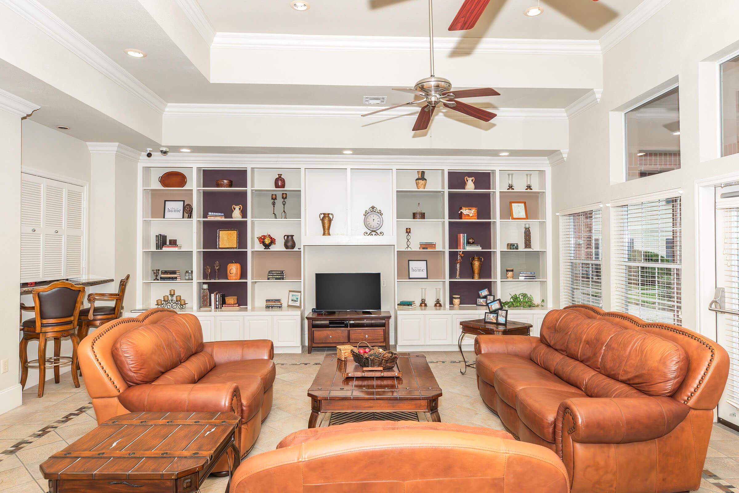 Spacious living room with high ceilings, featuring two brown leather sofas, a wooden coffee table, and a television on a white cabinet. Shelves display decorative items and plants. A dining area with wooden bar stools is visible in the background, with large windows letting in natural light.