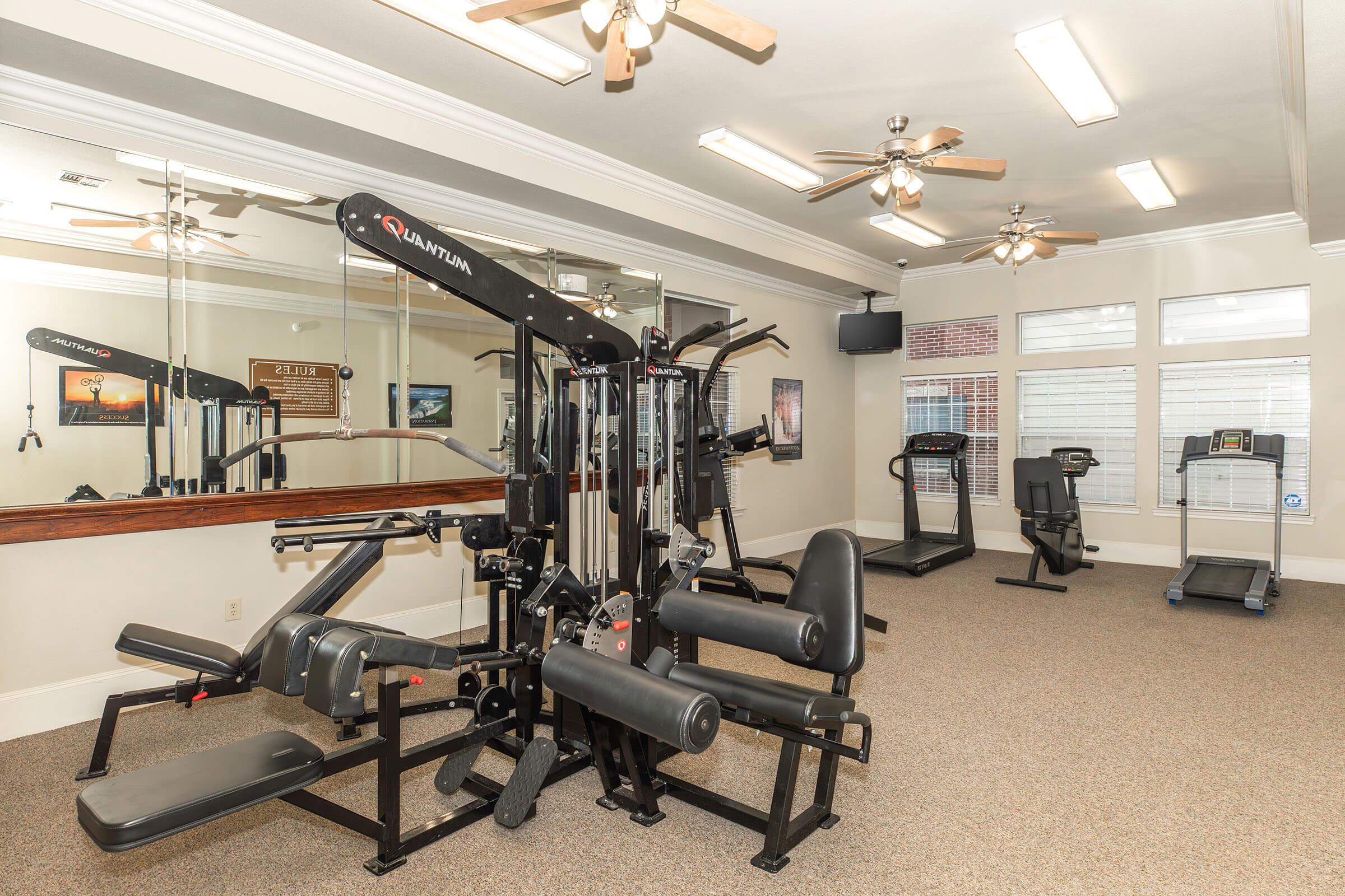 A well-equipped gym featuring strength training machines, a treadmill, and an exercise bike, all set in a bright room with mirrors and ceiling fans. The space has a clean, modern aesthetic with light-colored walls and carpeting, ideal for fitness enthusiasts.