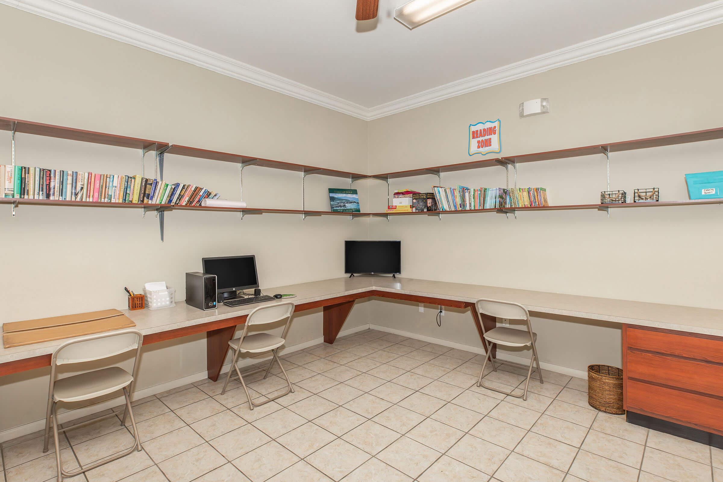A tidy office space featuring two workstations with computers on each desk. Shelves mounted on the walls hold a variety of books and decorative items. The floor is tiled, and there is a light-colored wall with a ceiling fan. A small basket is placed in the corner, giving the room a clean and organized look.