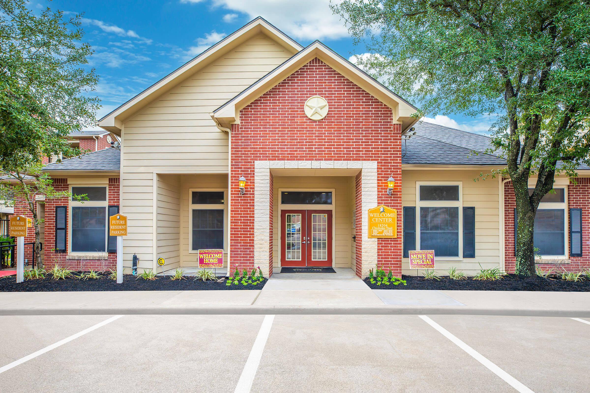 A well-maintained brick building with a welcoming entrance, featuring large windows and a red front door. Lush greenery is present in the landscaping, with signs displaying welcoming messages. The building is set against a clear blue sky, creating an inviting atmosphere.