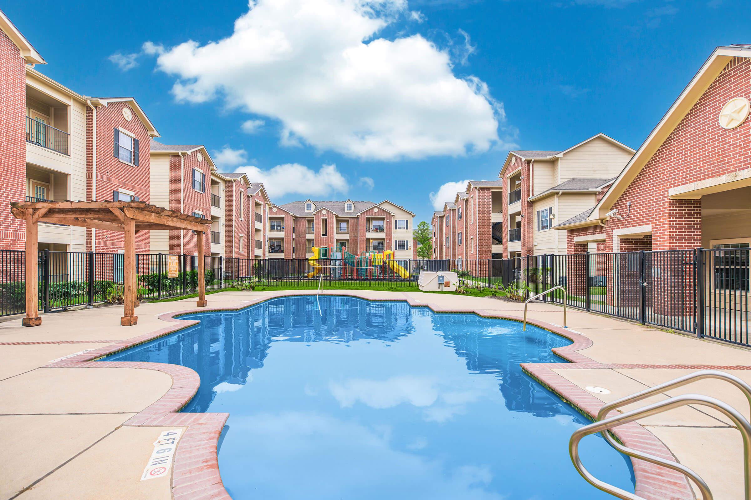 View of a residential apartment complex featuring a clear blue swimming pool surrounded by a deck. The pool is bordered by a black fence, with green landscaping and playground equipment visible in the background. The sky is bright with fluffy clouds, highlighting a sunny day.