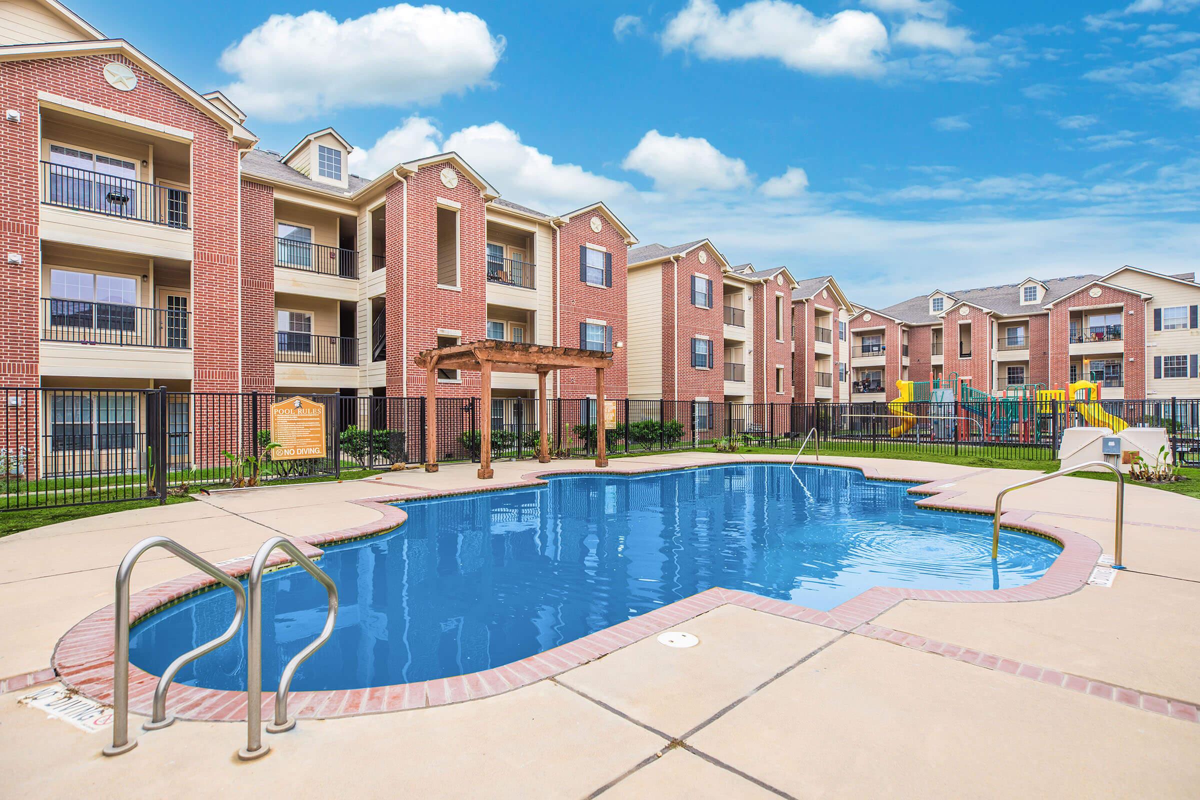 A view of a residential apartment complex featuring a swimming pool with a surrounding deck, lounge chairs, and a shaded pergola. In the background, multiple brick buildings with balconies are visible, along with a playground area. The sky is partly cloudy, adding to the inviting atmosphere.