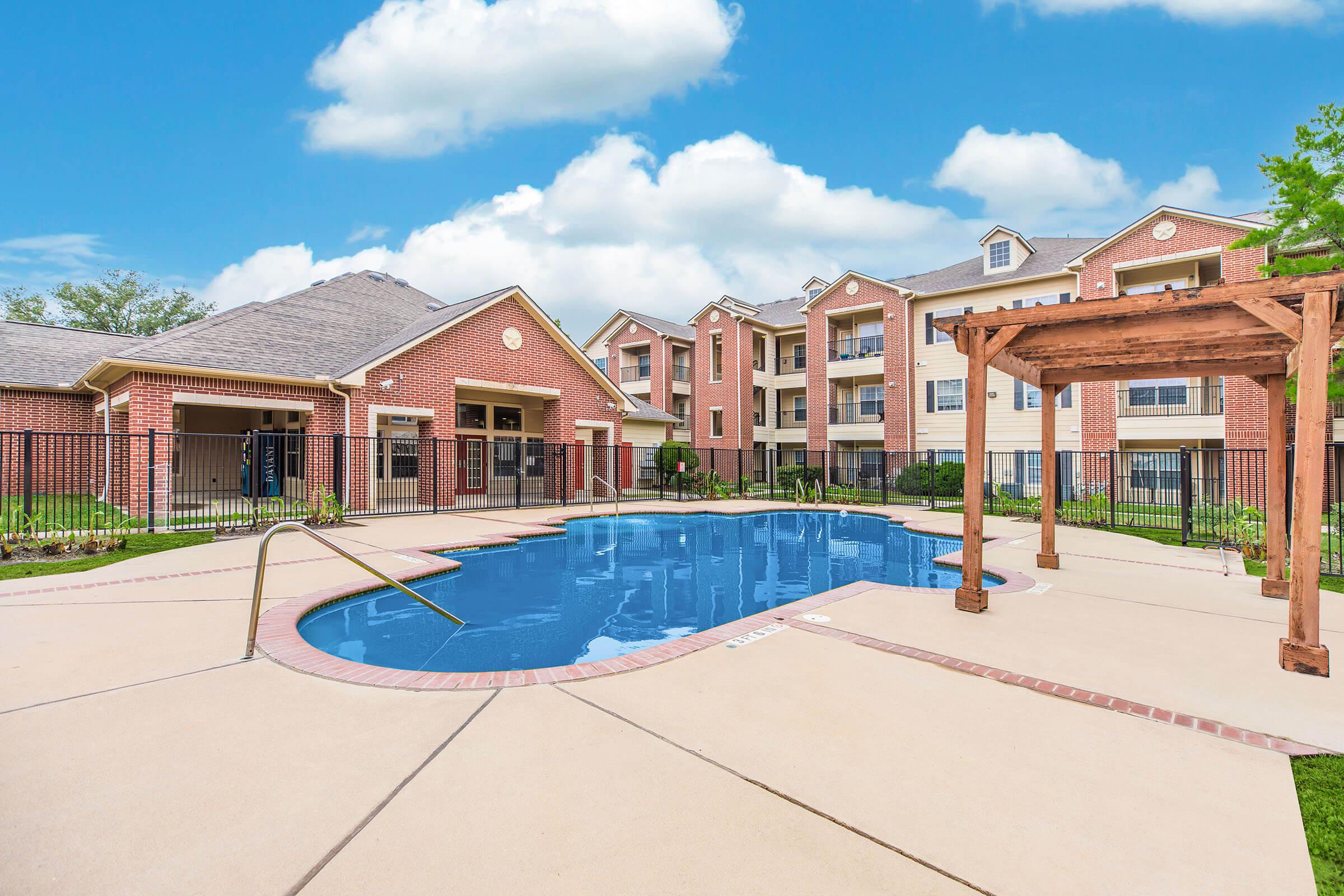 A large outdoor swimming pool surrounded by a concrete deck, with a wooden pergola and lounge area nearby. In the background, there are modern apartment buildings with red brick and beige siding, set against a clear blue sky with fluffy white clouds.