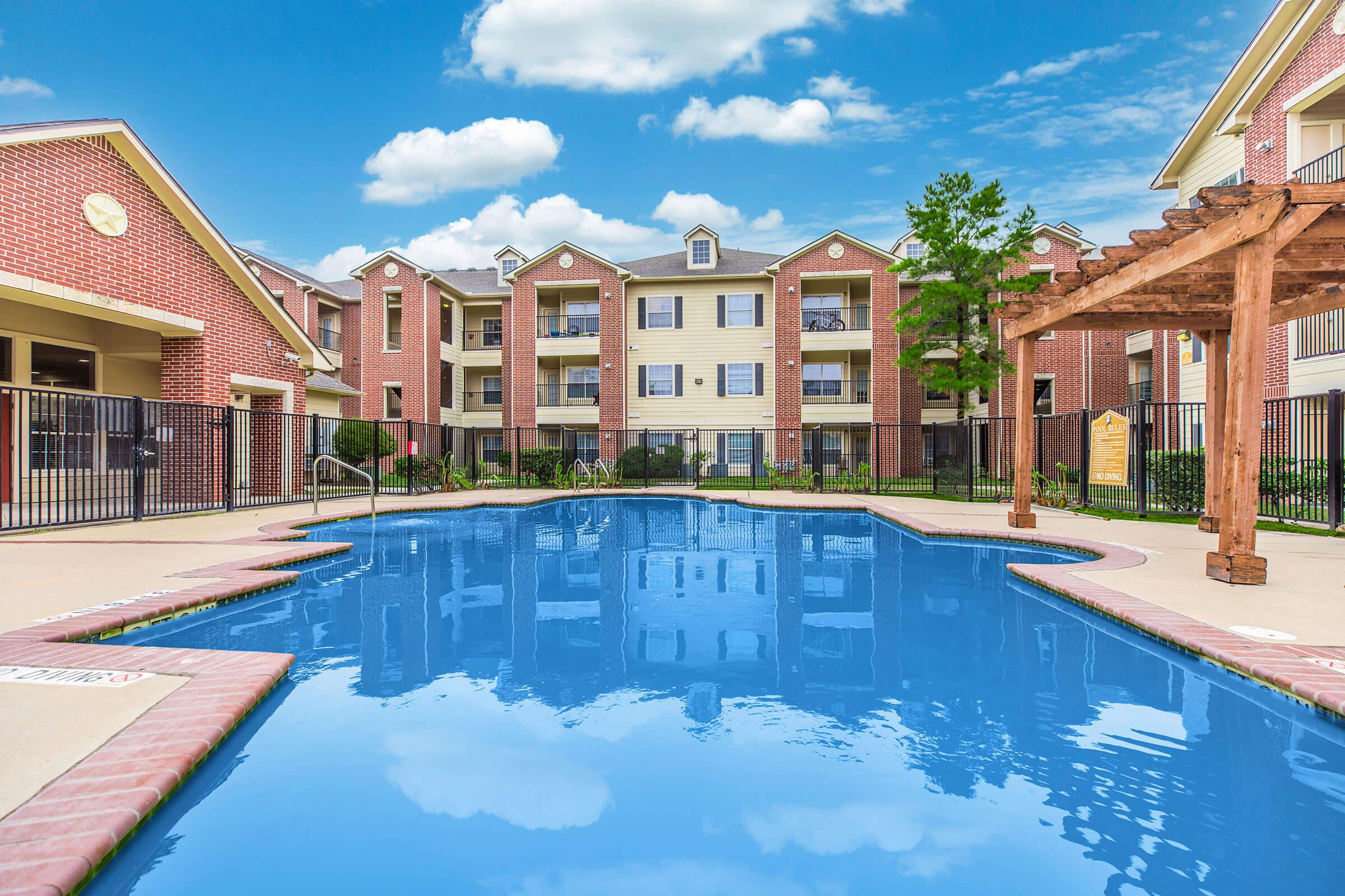 A clear blue swimming pool surrounded by an apartment complex. The pool features a shallow entry area and is framed by vibrant green grass and landscaped areas. The buildings are multi-story with brick and siding exteriors, and the sky above is bright with fluffy white clouds.