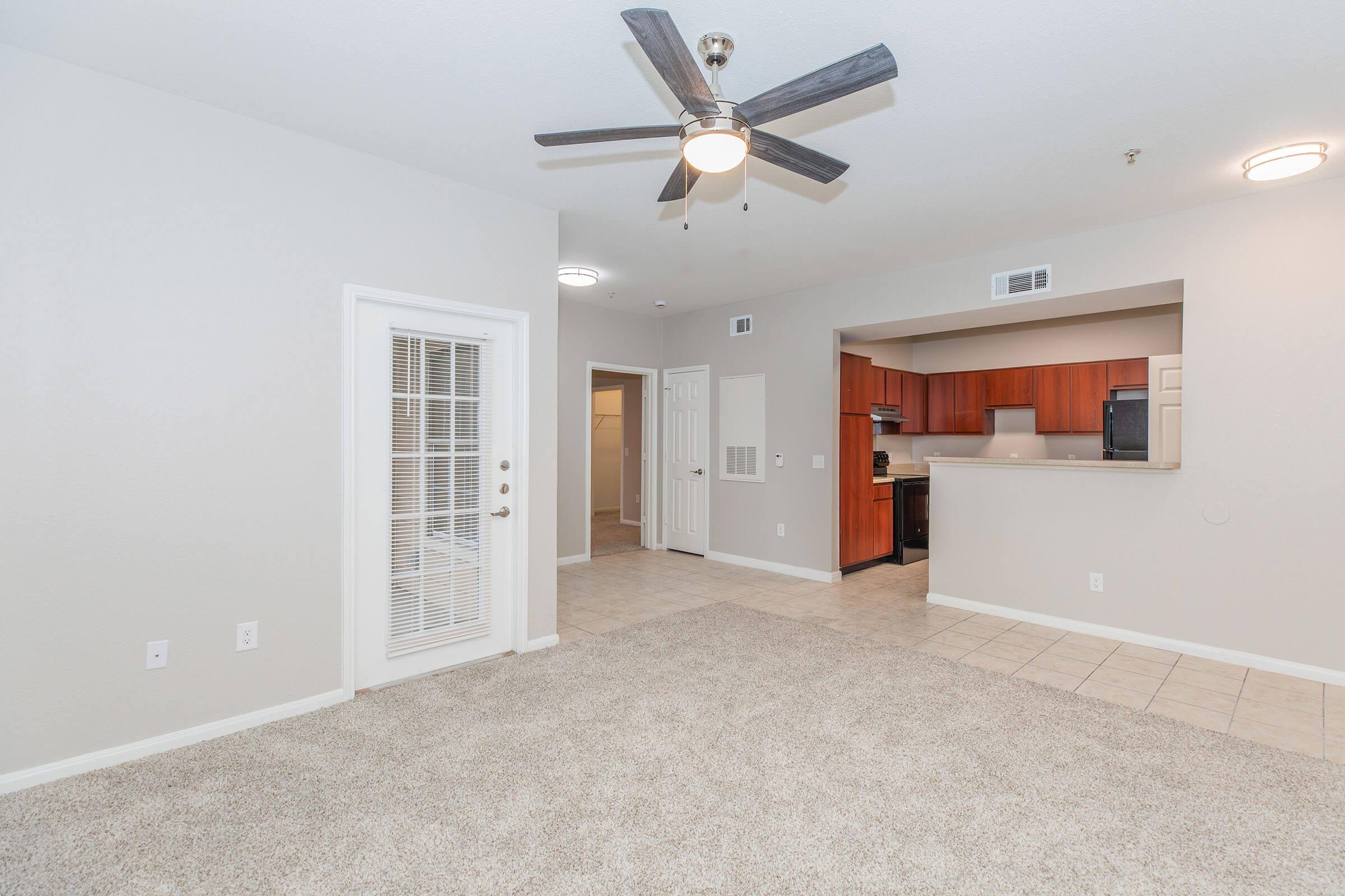 A spacious living area with light-colored walls and a ceiling fan. The floor is carpeted, and there's a set of double French doors leading to another room. In the background, an open-concept kitchen features wooden cabinetry and modern appliances, with tile flooring visible.