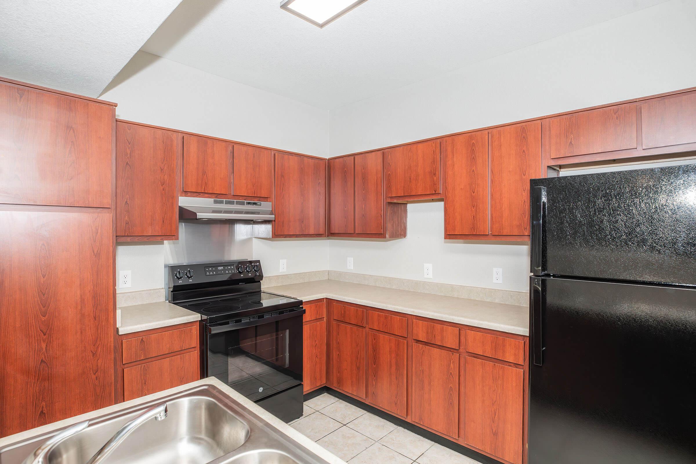 Modern kitchen featuring wooden cabinets, stainless steel appliances including an oven and microwave, and a black refrigerator. The countertop is light-colored, and the tile floor is neutral-toned. There are no items on the countertops or inside the cabinets, giving a clean and uncluttered appearance.