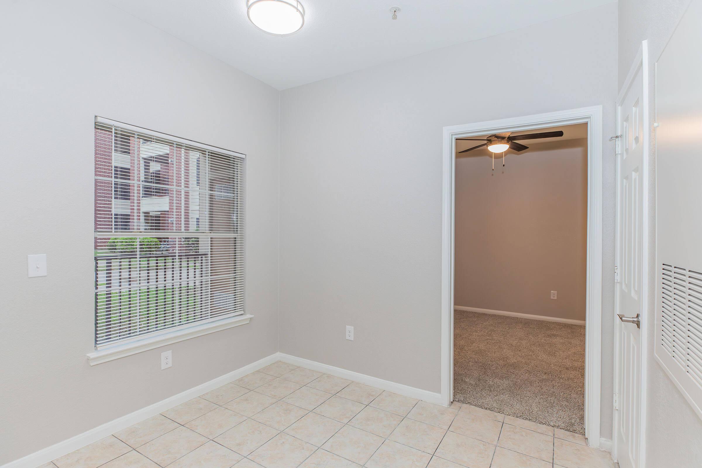 A small, empty room with light gray walls and tiled flooring. A window with blinds lets in natural light, and a door leads to another room with beige carpet and a ceiling fan. The overall ambiance is bright and minimalistic.