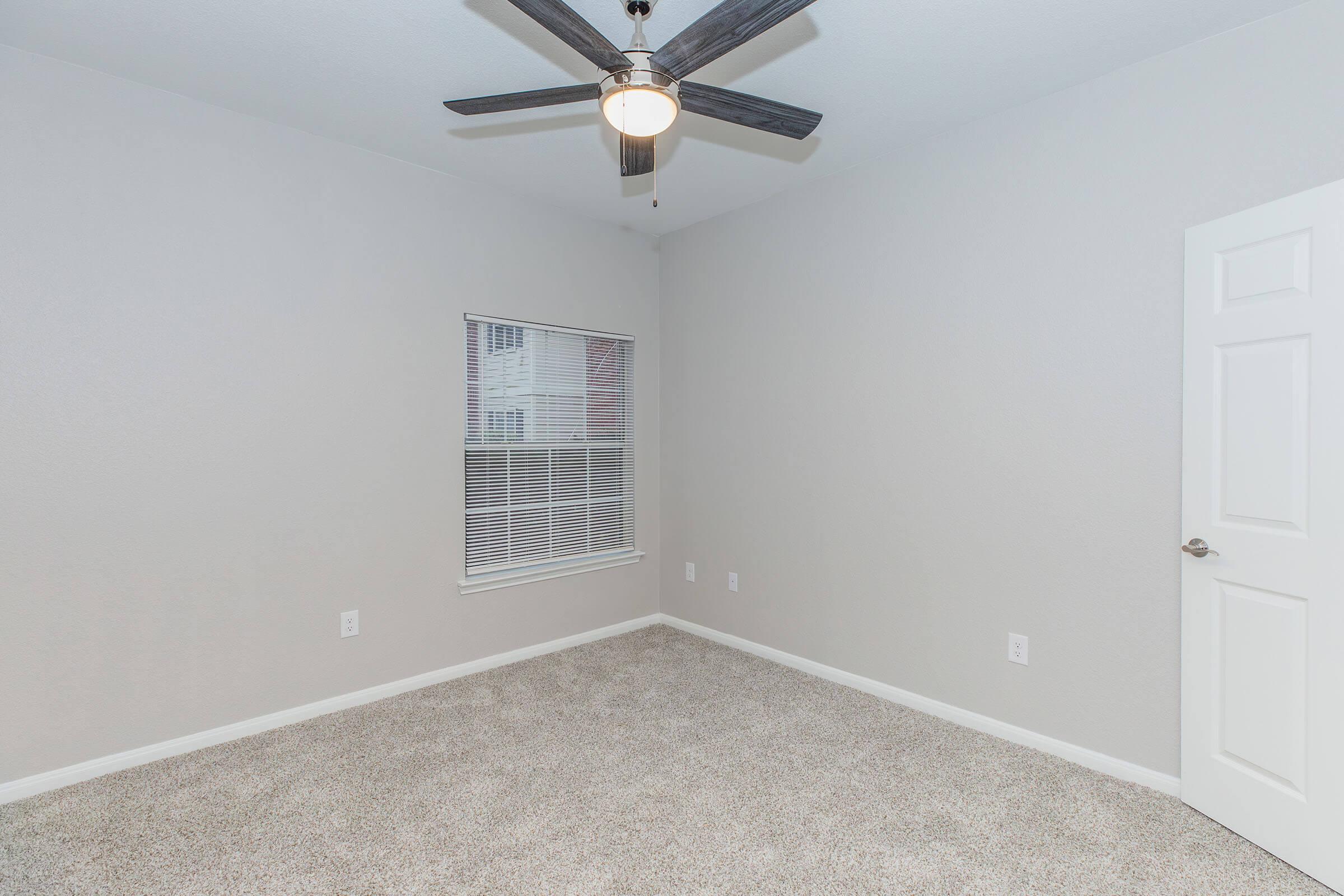 A clean, empty bedroom featuring light gray walls and a ceiling fan. The floor is covered with beige carpet, and there is a window with blinds on one wall, allowing natural light to enter. A white door is visible on the right side of the image.