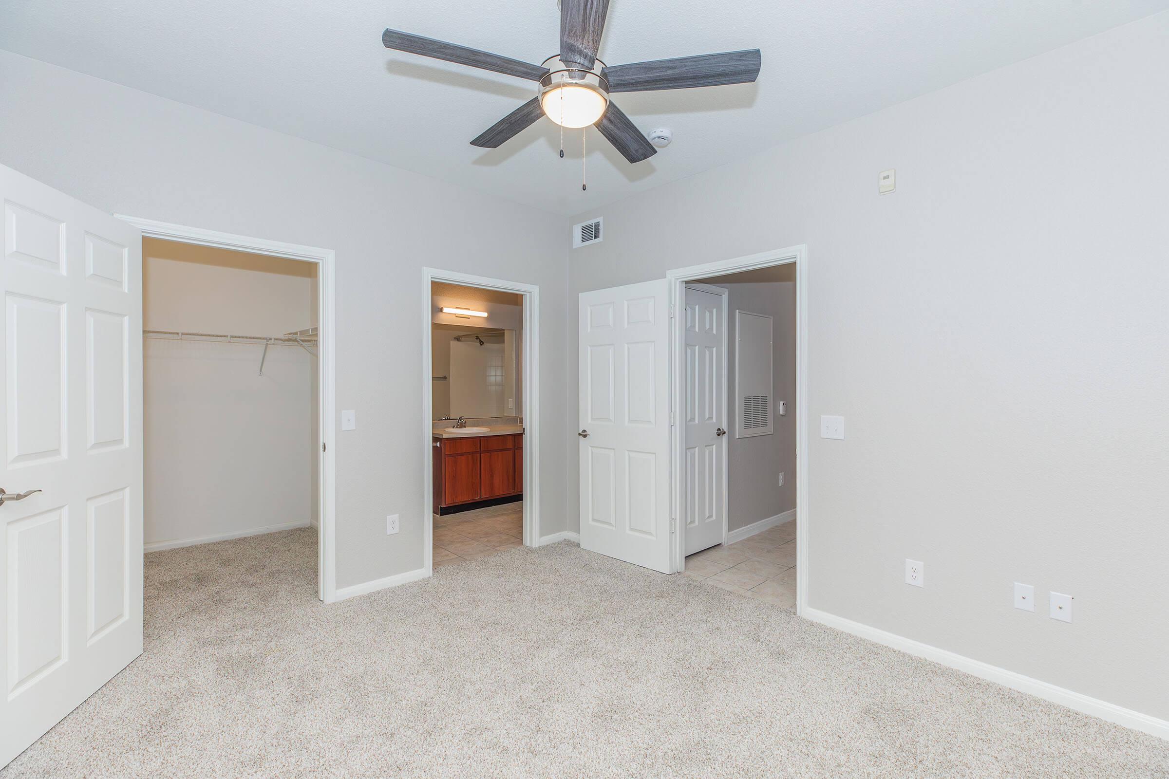 A well-lit, freshly painted room featuring beige walls and plush carpeting. It includes two open doorways leading to a closet and a bathroom, and a ceiling fan with wooden blades hangs above. The room is empty, creating a spacious feel, ideal for various furniture arrangements.