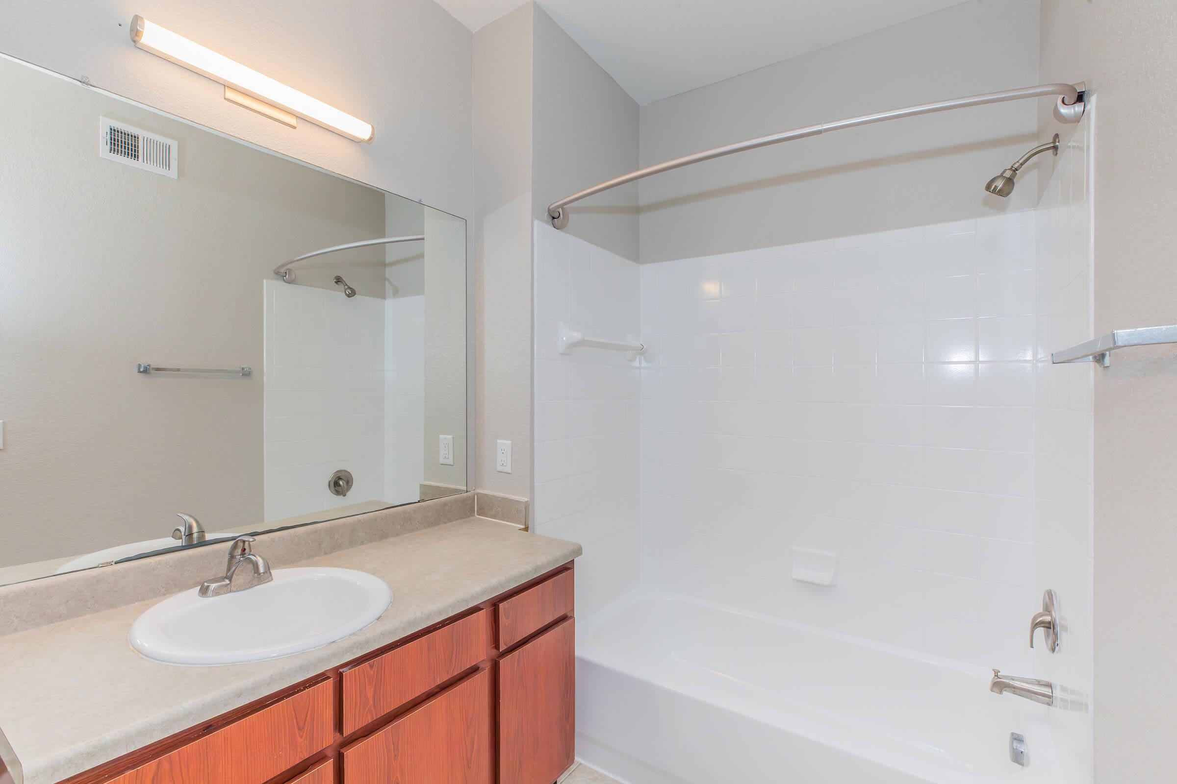 A clean, modern bathroom featuring a white bathtub with a shower curtain rod, a large mirror above a beige countertop, and wooden cabinets. The walls are painted in a light color, and there is soft lighting from a fixture above the mirror. The space is tidy and well-maintained.