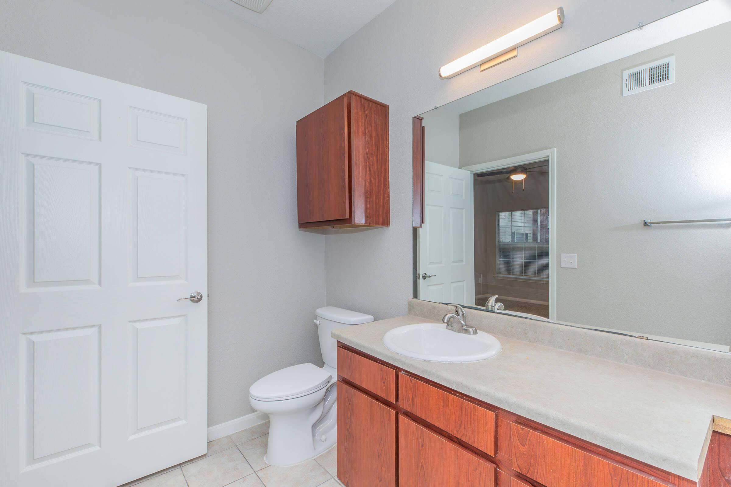 A clean, modern bathroom featuring a double sink vanity with wooden cabinets, a large mirror above, and a white toilet. The walls are light gray, and there's a door leading to another room. The floor is tiled, and there is a towel bar on the wall. Natural light enhances the space.