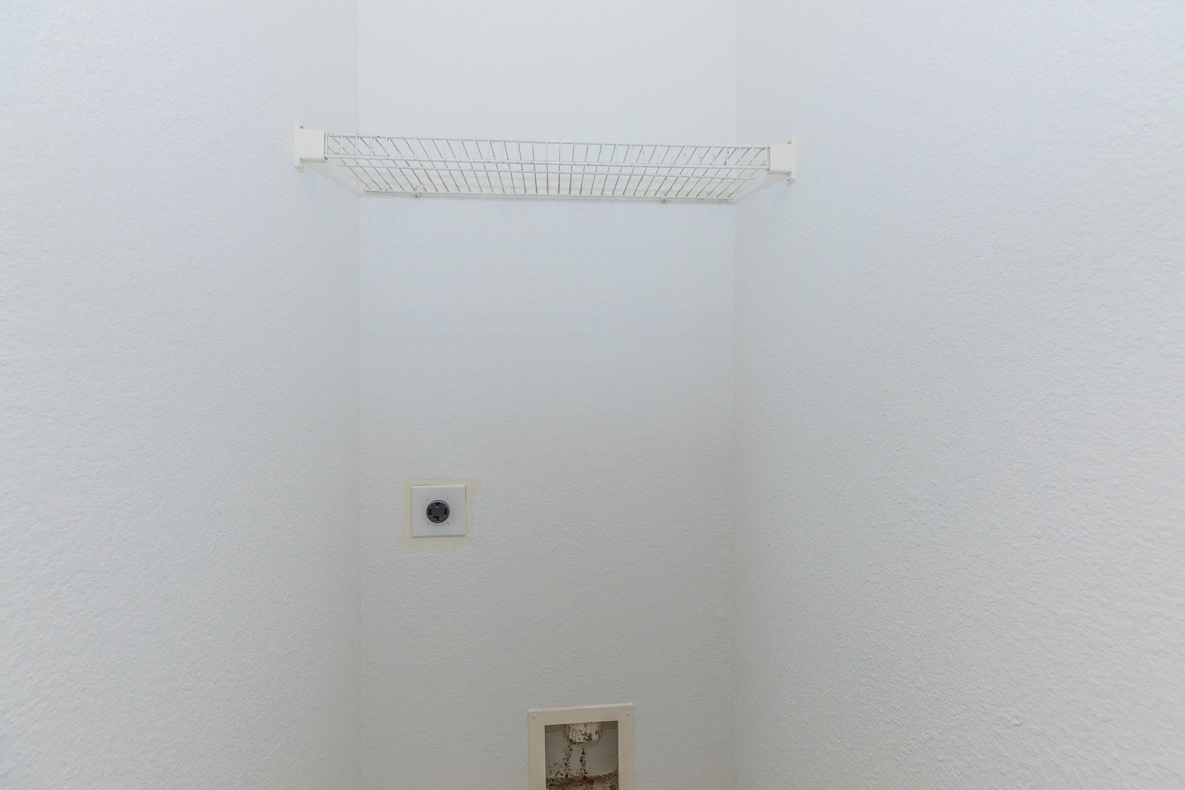 Empty closet space featuring plain white walls. A wire shelf is mounted several feet above the floor, and a small electrical outlet is visible to the left side of the wall. The floor appears unfinished, with a plumbing fixture partially visible near the bottom. Overall, the space looks minimalist and unadorned.