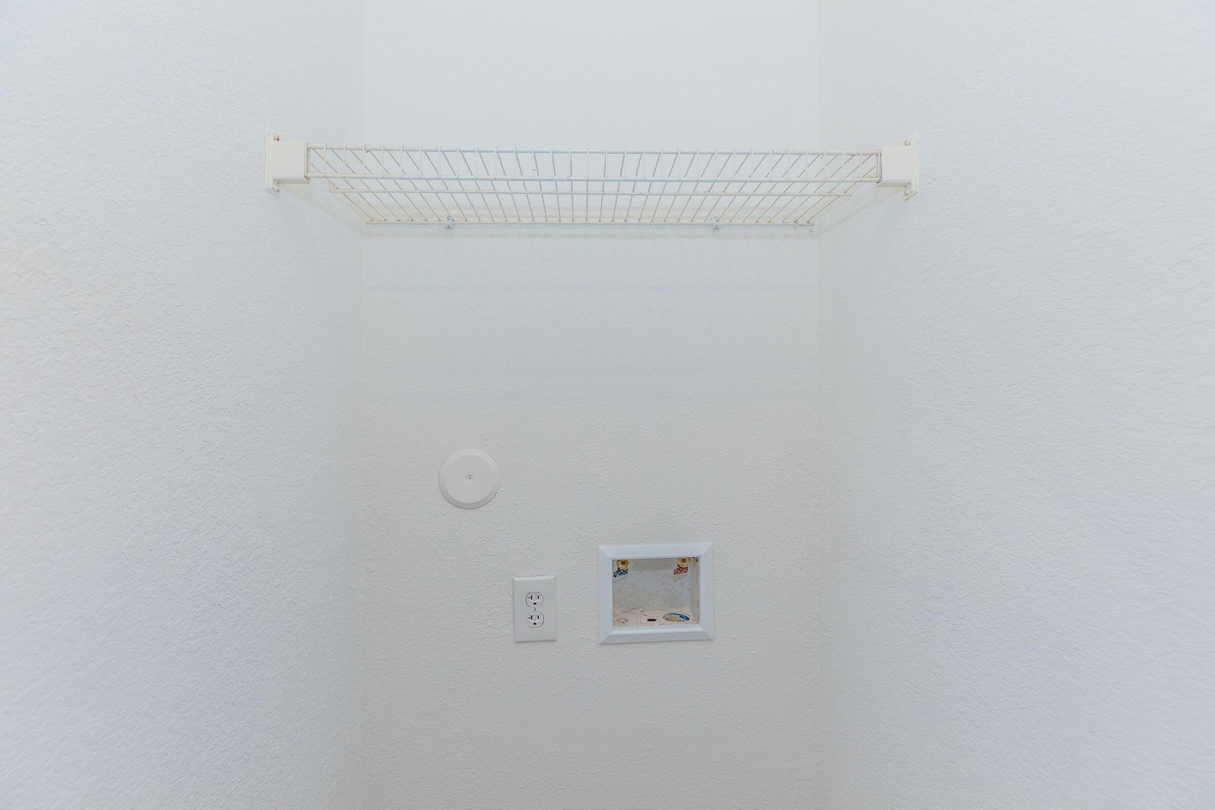 Empty white wall featuring a wire shelf mounted near the top. Below the shelf, there is an electric outlet and a small, framed opening in the wall. The overall space has a clean and minimalist appearance.