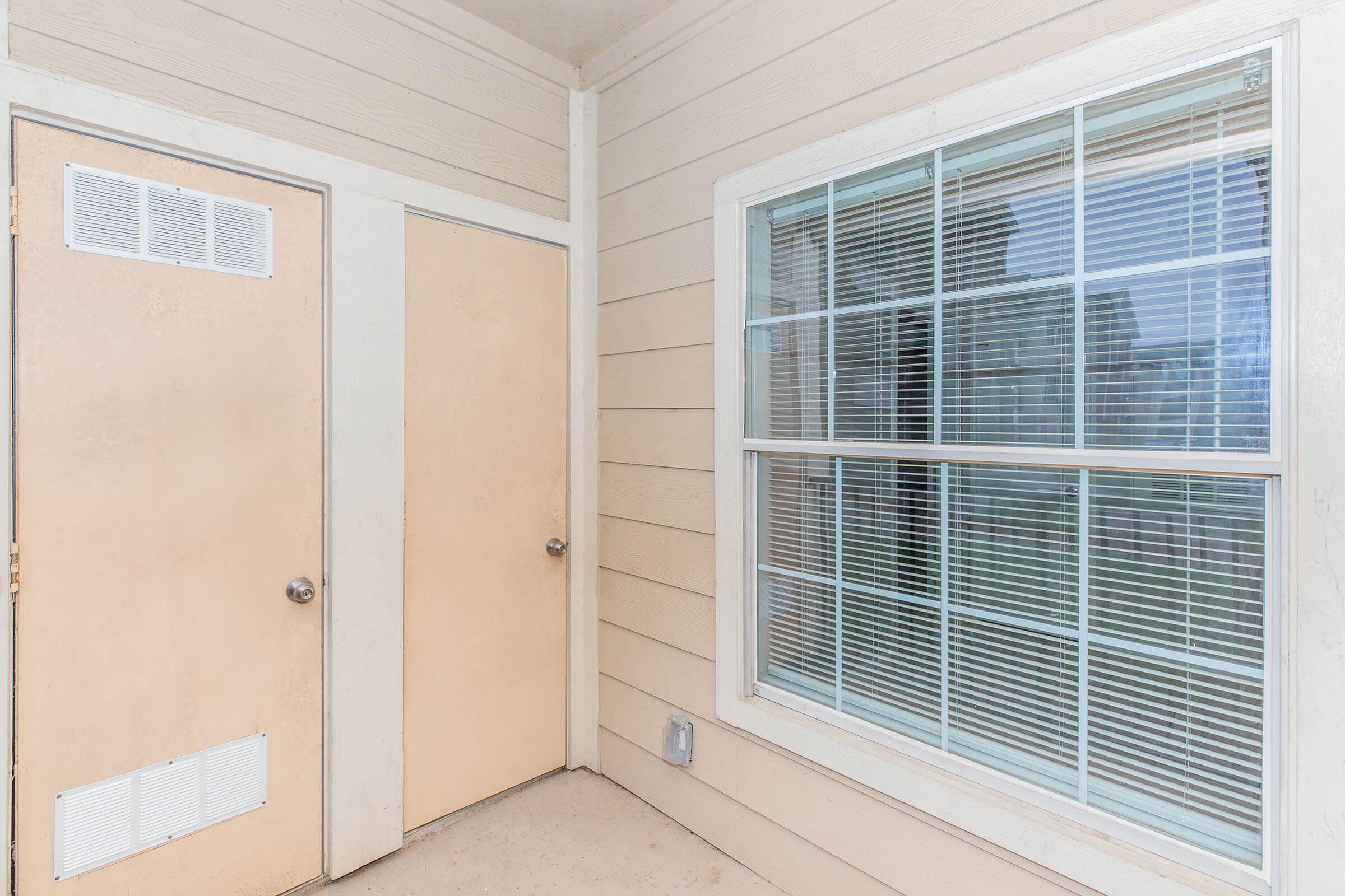 A corner of an exterior wall featuring a window with vertical blinds and two closed doors, set against a neutral-colored siding. The window reflects the surroundings, while the doors have a simple design. The area appears well-maintained and invites natural light.