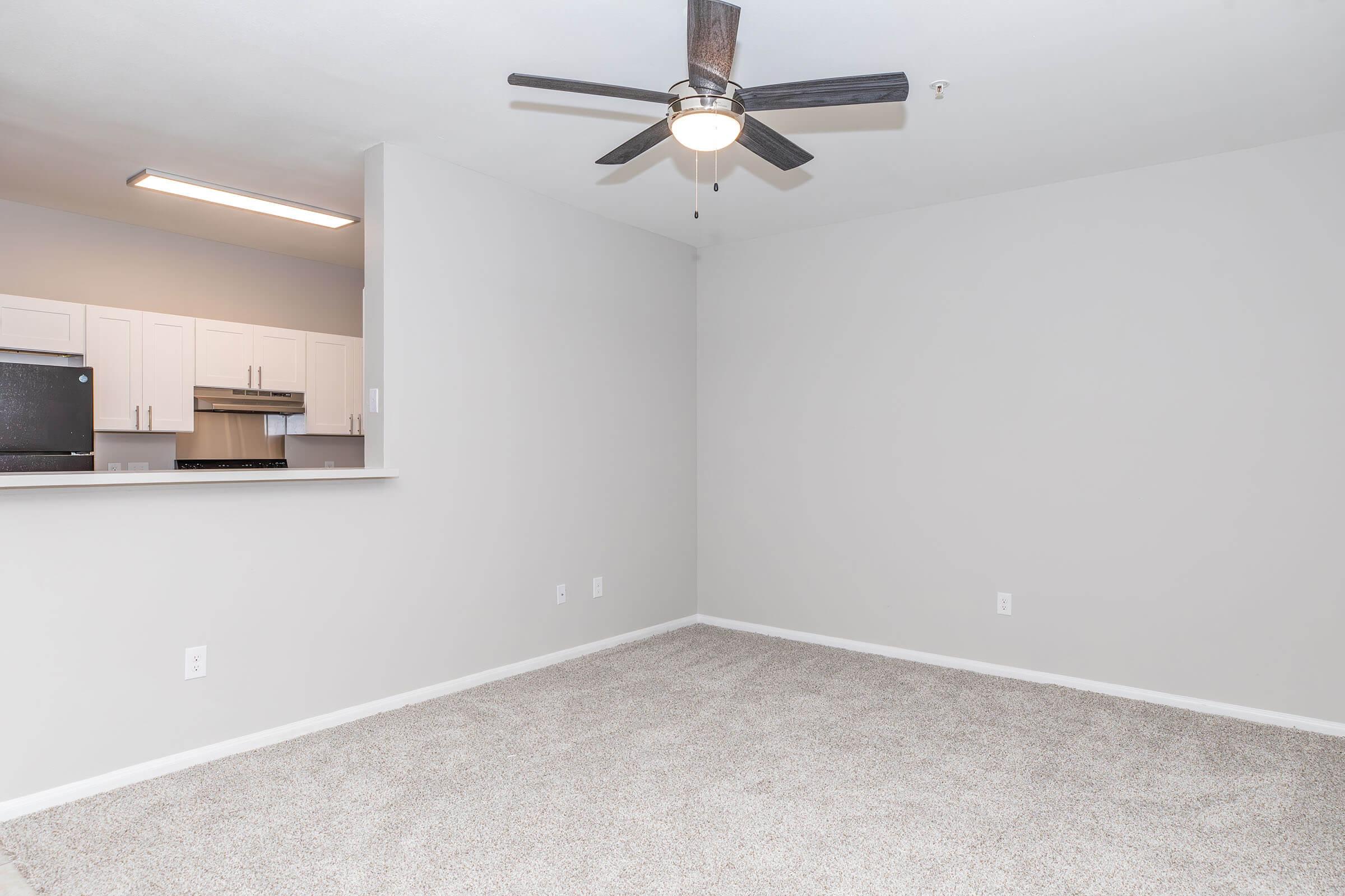 An empty living room with light gray walls and a ceiling fan. The space features beige carpet and an open area leading to a kitchen with white cabinets and a dark countertop, visible in the background. Natural light fills the room, contributing to a bright, airy feel.