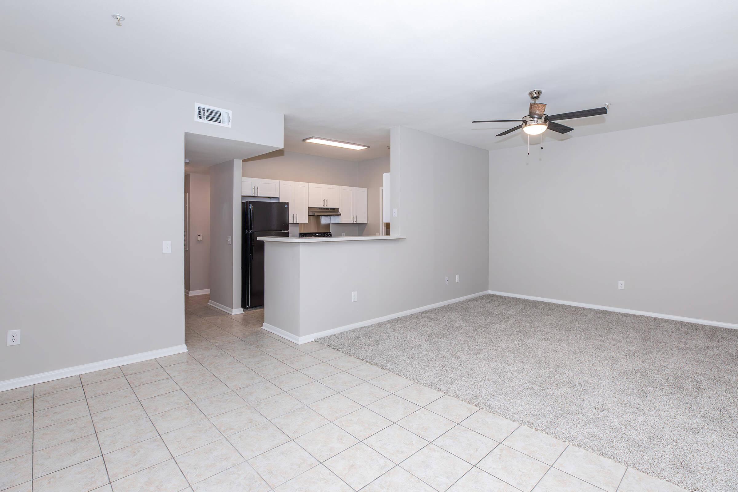 A spacious, empty living room featuring light gray walls, tiled flooring, and a large carpeted area. A ceiling fan is mounted above, and an open kitchen area with modern appliances is visible in the background. The room is well-lit and has ample natural light.