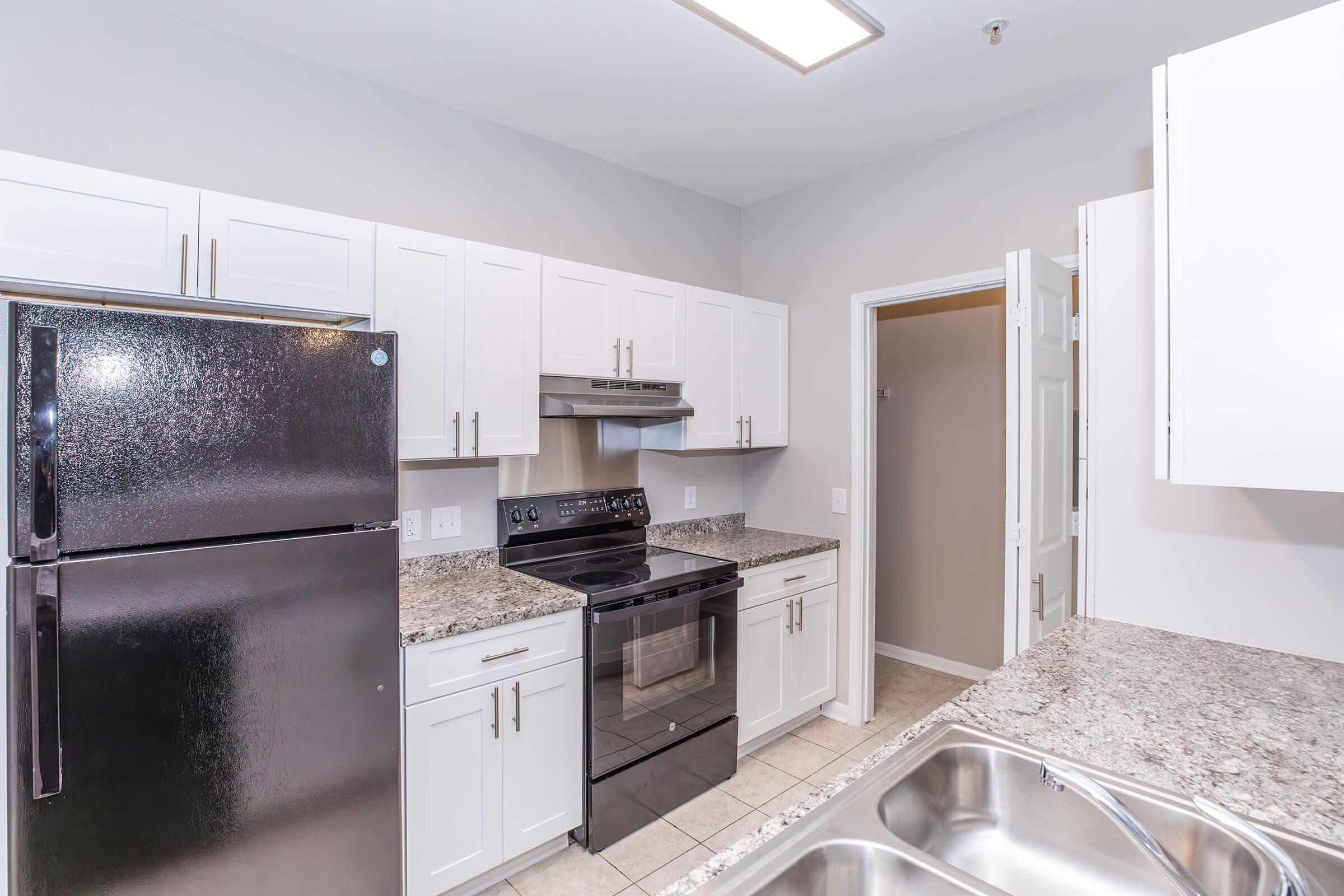 Modern kitchen featuring white cabinets, black refrigerator, stainless steel oven, and granite countertops. The layout includes a double sink, and the space is well-lit with overhead lighting. A door leads to another room or pantry, adding functionality to the design.