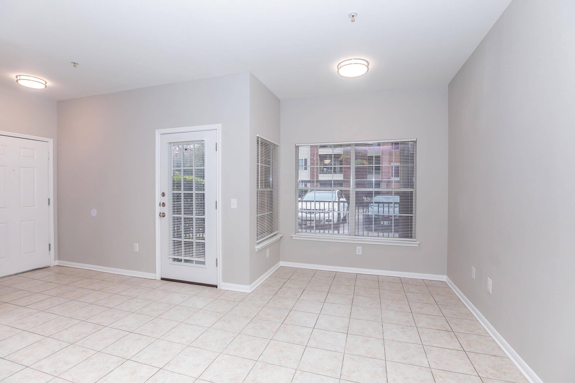 A spacious, empty room with light gray walls and tiled flooring. It features a door leading outside and a window with blinds, providing natural light. The overall atmosphere is bright and open, suitable for various interior decorating options.