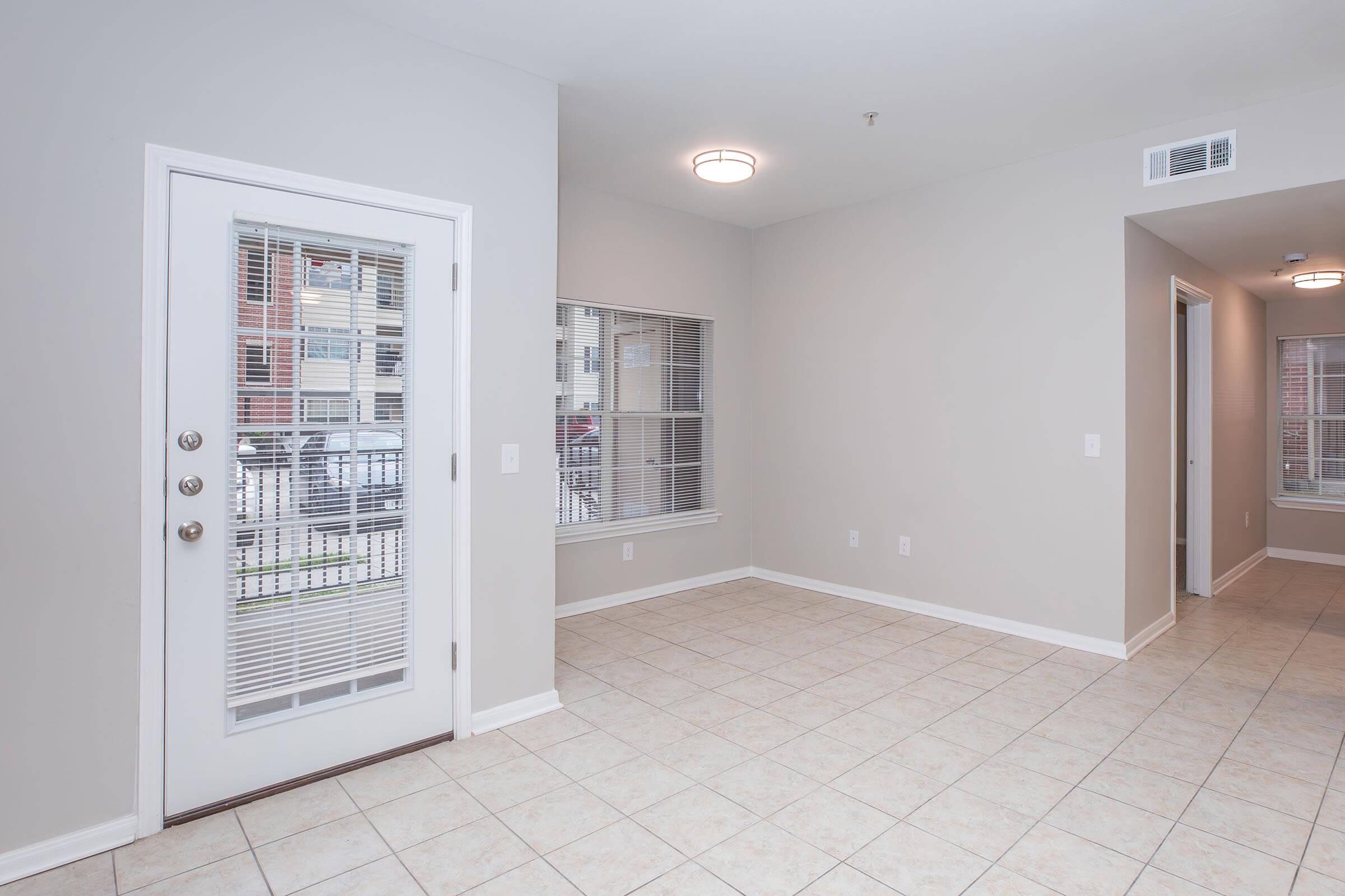 A spacious, empty room with light-colored walls and tiled flooring. A door with glass panels leads outside, and there are two windows with blinds letting in natural light. The room is well-lit with a ceiling fixture, showcasing a modern and clean interior design.
