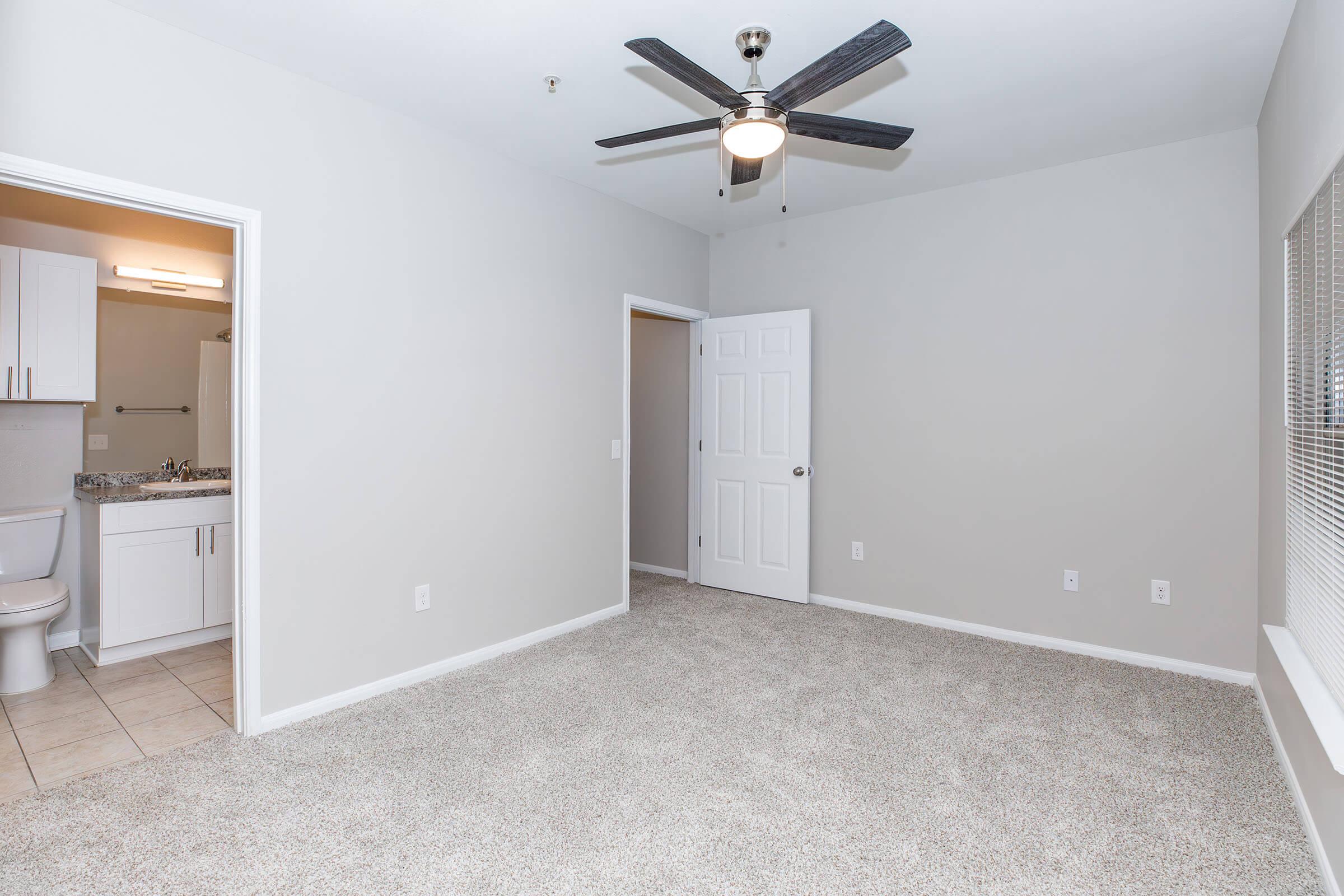 A spacious living area with light gray walls and plush carpet, featuring a ceiling fan. To the left, there is an open doorway leading to a bathroom with a modern sink and toilet. Natural light enters through a window, enhancing the room's bright and airy feel.