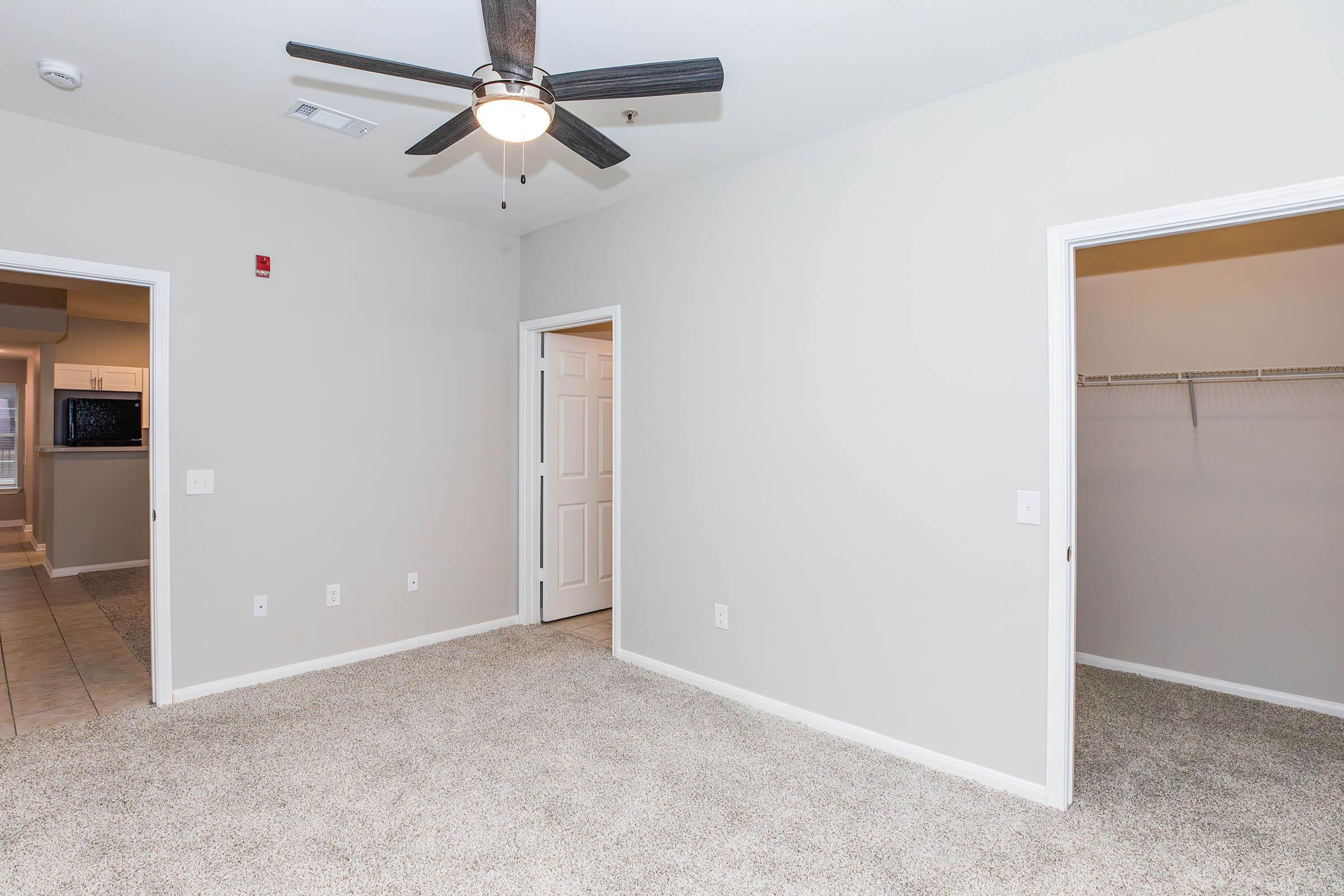 A well-lit, empty room with light-colored walls and carpet. It features a ceiling fan, two doorways leading to adjacent rooms, and a closet with shelving. The room appears spacious and organized, with minimal furniture or decor, creating a clean and inviting atmosphere.