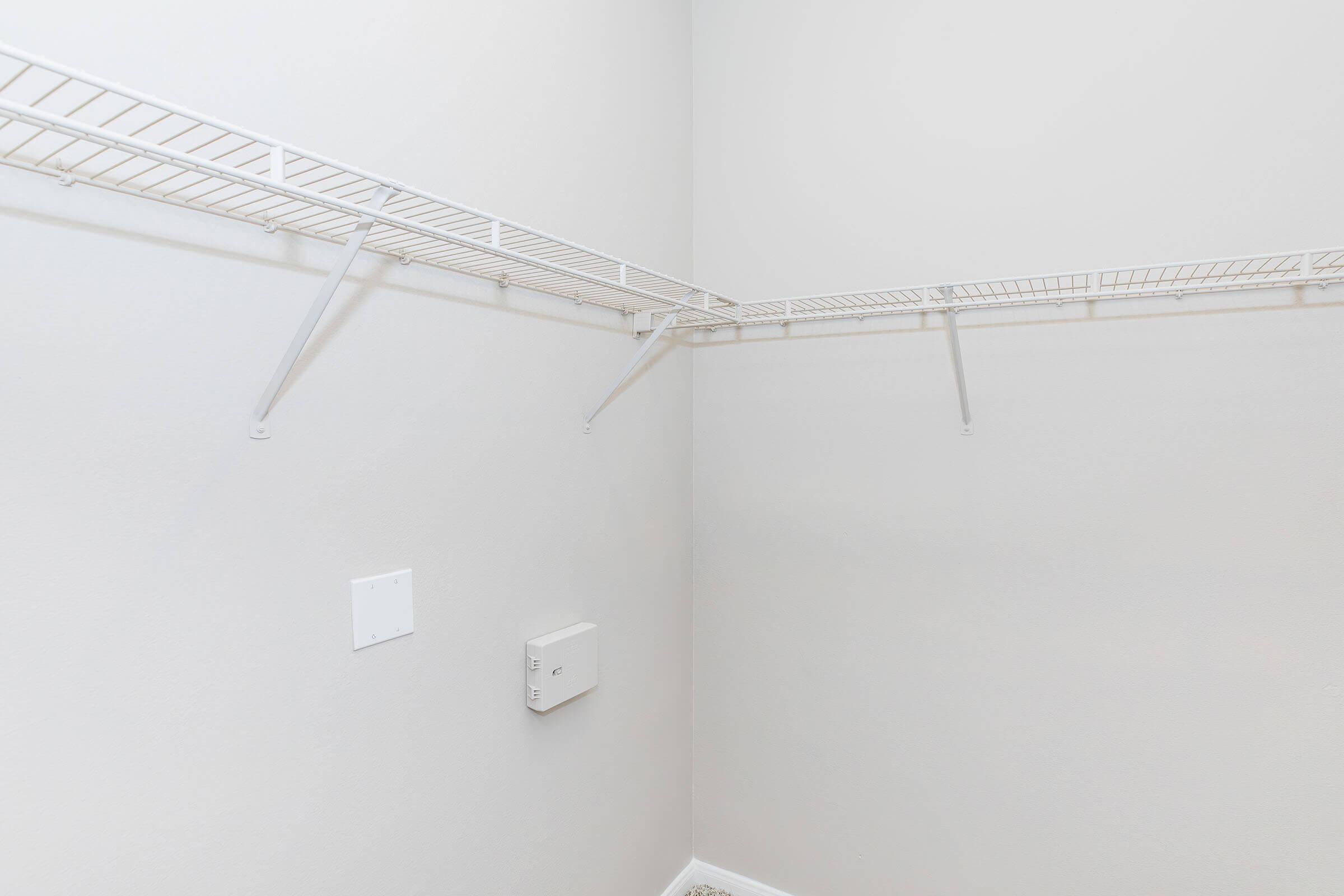 An empty closet with light gray walls and a wire shelving unit mounted on the back wall. The shelving is positioned in a corner, with no items or decorations visible. A small electrical box is mounted on the wall below the shelves. The floor is not visible in the image.