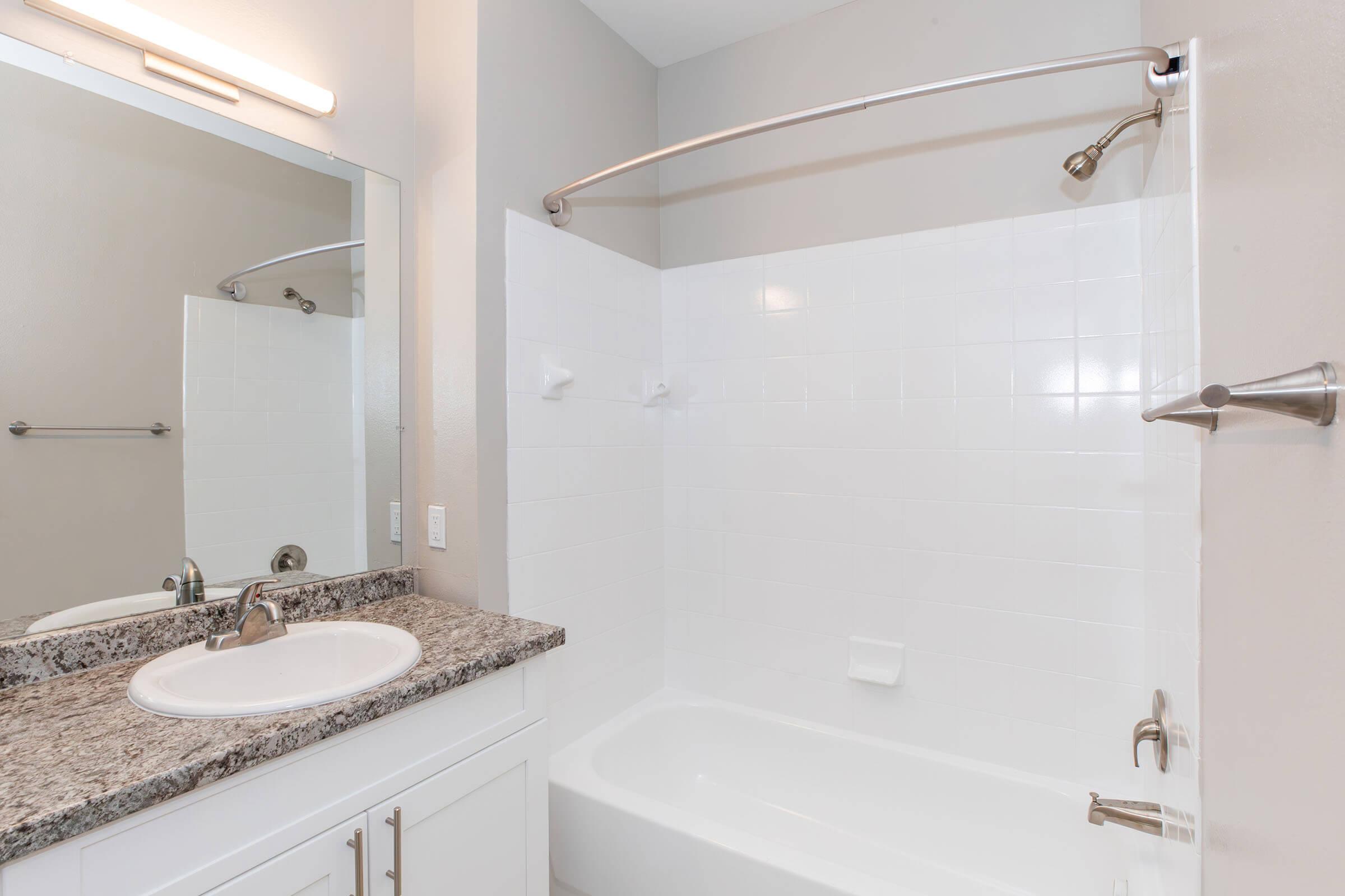 A modern, clean bathroom featuring a white shower/tub combo, a granite countertop with a sink, a wall-mounted mirror, and neutral-colored walls. The design is minimalistic with chrome fixtures, providing a bright and spacious appearance.