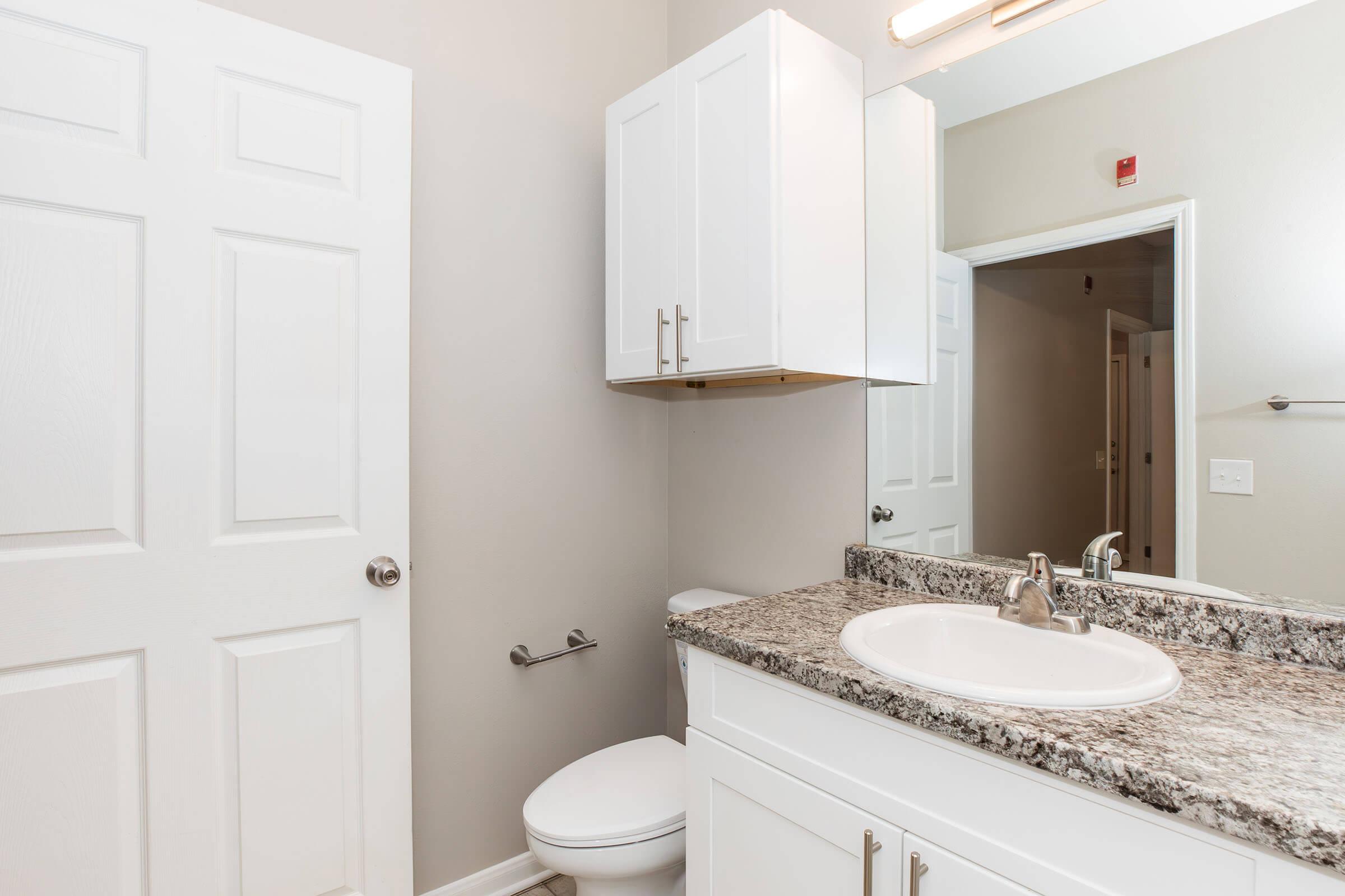 A modern bathroom featuring light-colored walls, a granite countertop sink area, a white cabinet above the sink, a toilet, and a large mirror. The door is partially open, and there is a towel rack on the wall, creating a clean and minimalistic aesthetic.