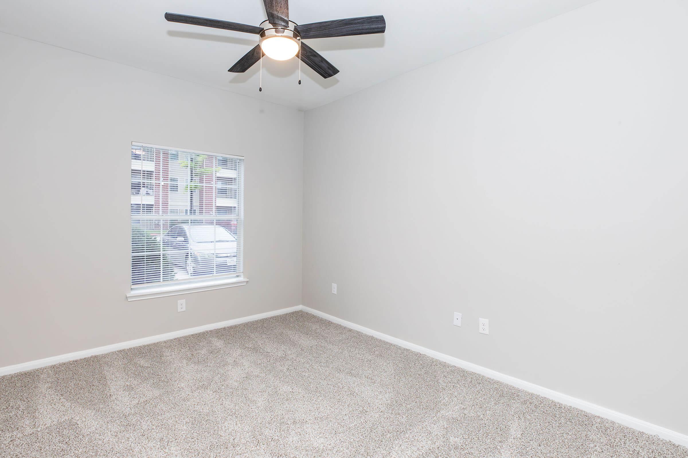 Empty room with light gray walls, carpeted floor, and a ceiling fan. A window with blinds lets in natural light, revealing a view of the exterior. The room is uncluttered, offering a blank space suitable for various uses such as a bedroom or office.