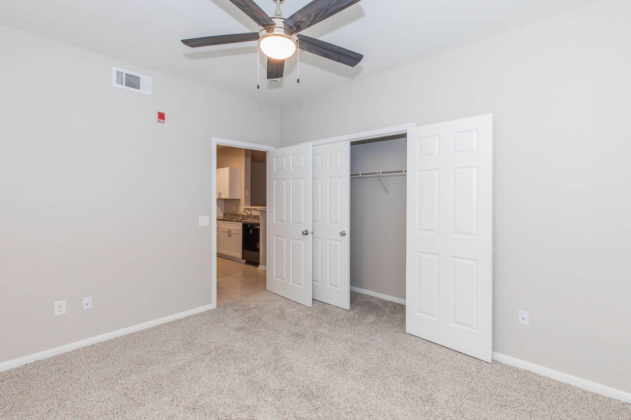 A well-lit room featuring neutral walls, a ceiling fan with wooden blades, and plush carpet. There are double doors leading to a closet on one side, while the other side opens to a kitchen. The space appears clean and modern, suitable for various uses like a bedroom or office.