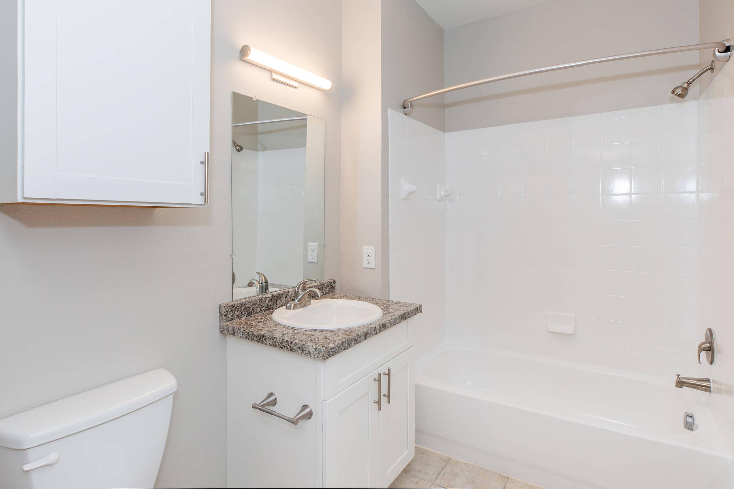 A clean and modern bathroom featuring a white tub with a shower rod, a single sink with a granite countertop, a large mirror above the sink, and a white cabinet. The walls are painted in a light color, and there is a small light fixture above the mirror. The flooring is neutral-colored tile.