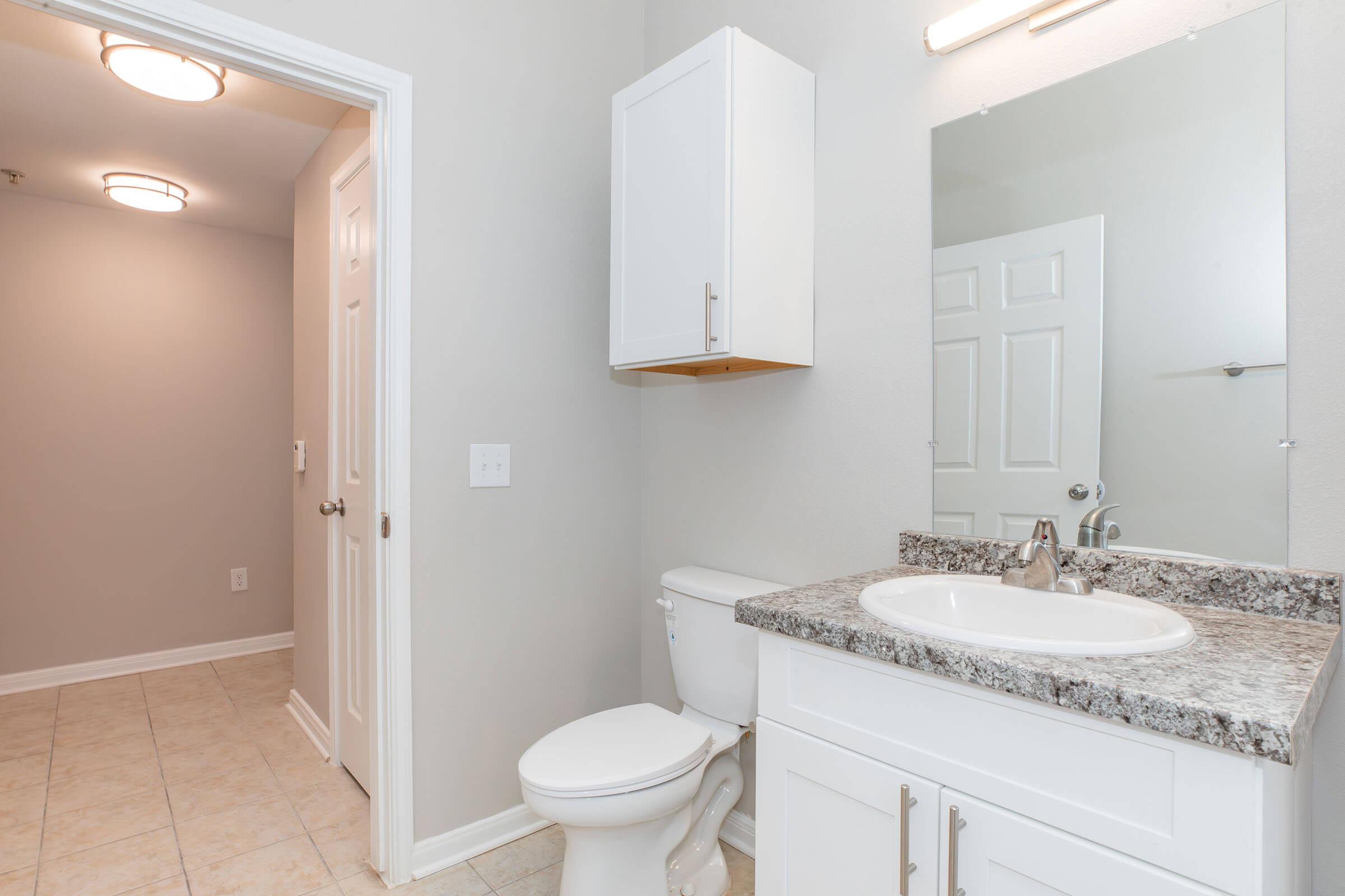 A clean and modern bathroom featuring a white vanity with a granite countertop, a round sink, a toilet, and a large mirror. The walls are painted light gray, and there is a doorway leading to a hallway. Ceiling lights illuminate the space, and the flooring is tiled.
