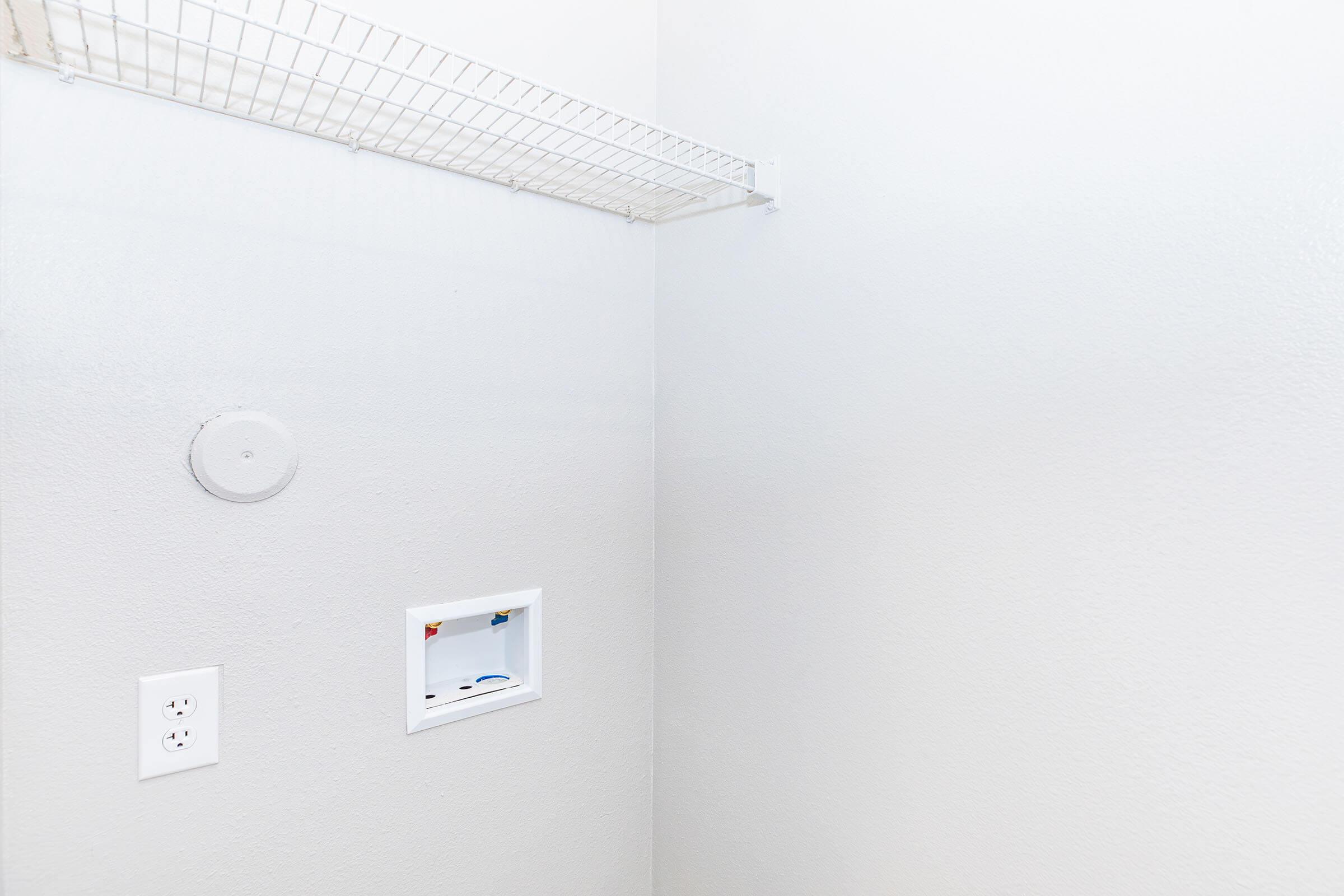 Empty room with light gray walls featuring a wire shelf mounted on the left side and an electrical outlet on the wall. In the corner, there is a small recessed area for a utility connection. The overall space is uncluttered and minimalistic, giving a modern appearance.