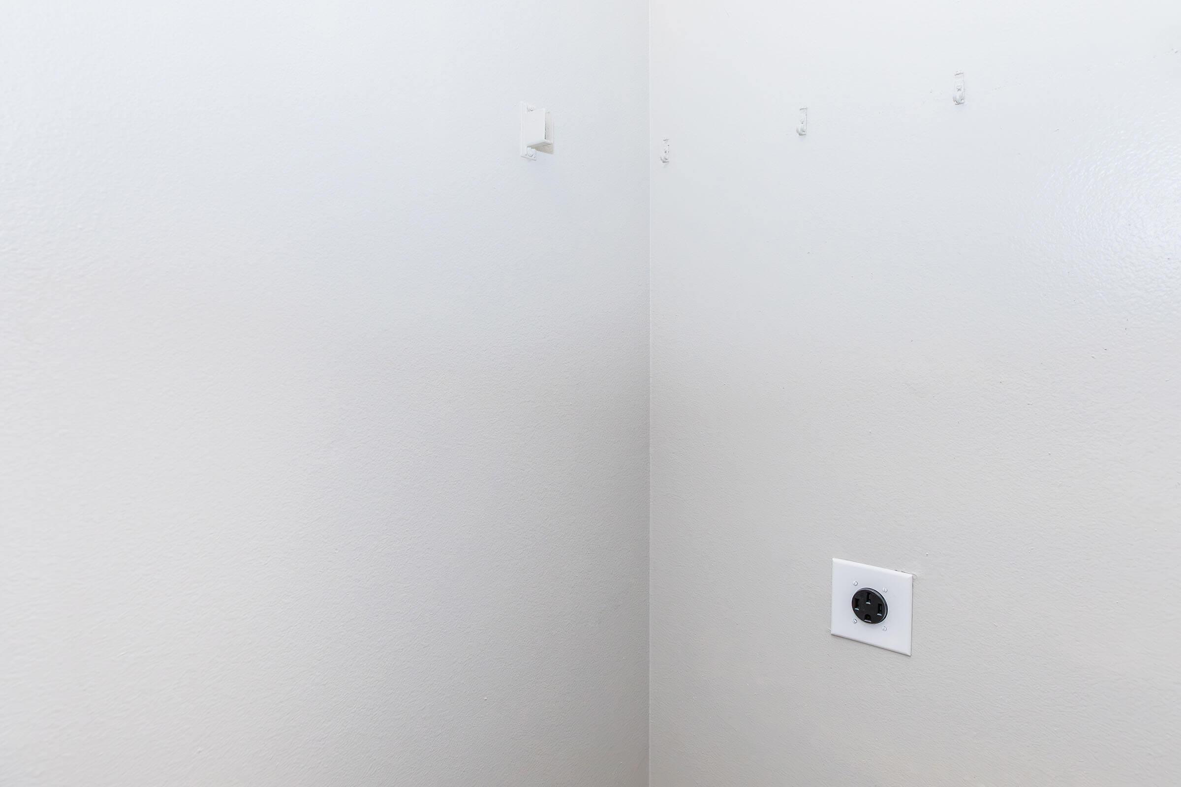 A plain white wall in a corner of a room, featuring two small, unpainted wall patches with no visible fixtures, and a power outlet on the lower right side. The overall setting is minimalistic and unadorned, emphasizing a clean and simple aesthetic.