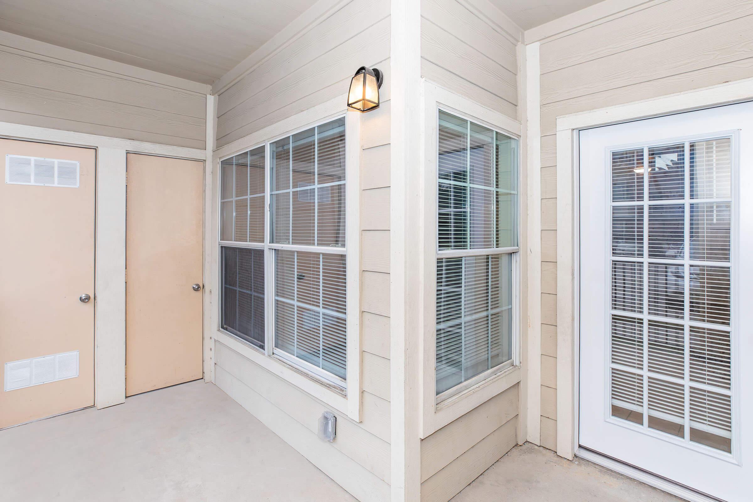 A corner view of a patio area in a residential building. The wall features large windows with blinds and a door with glass panes. There's a simple light fixture installed above the windows. The area has a neutral color scheme with beige siding and a concrete floor.